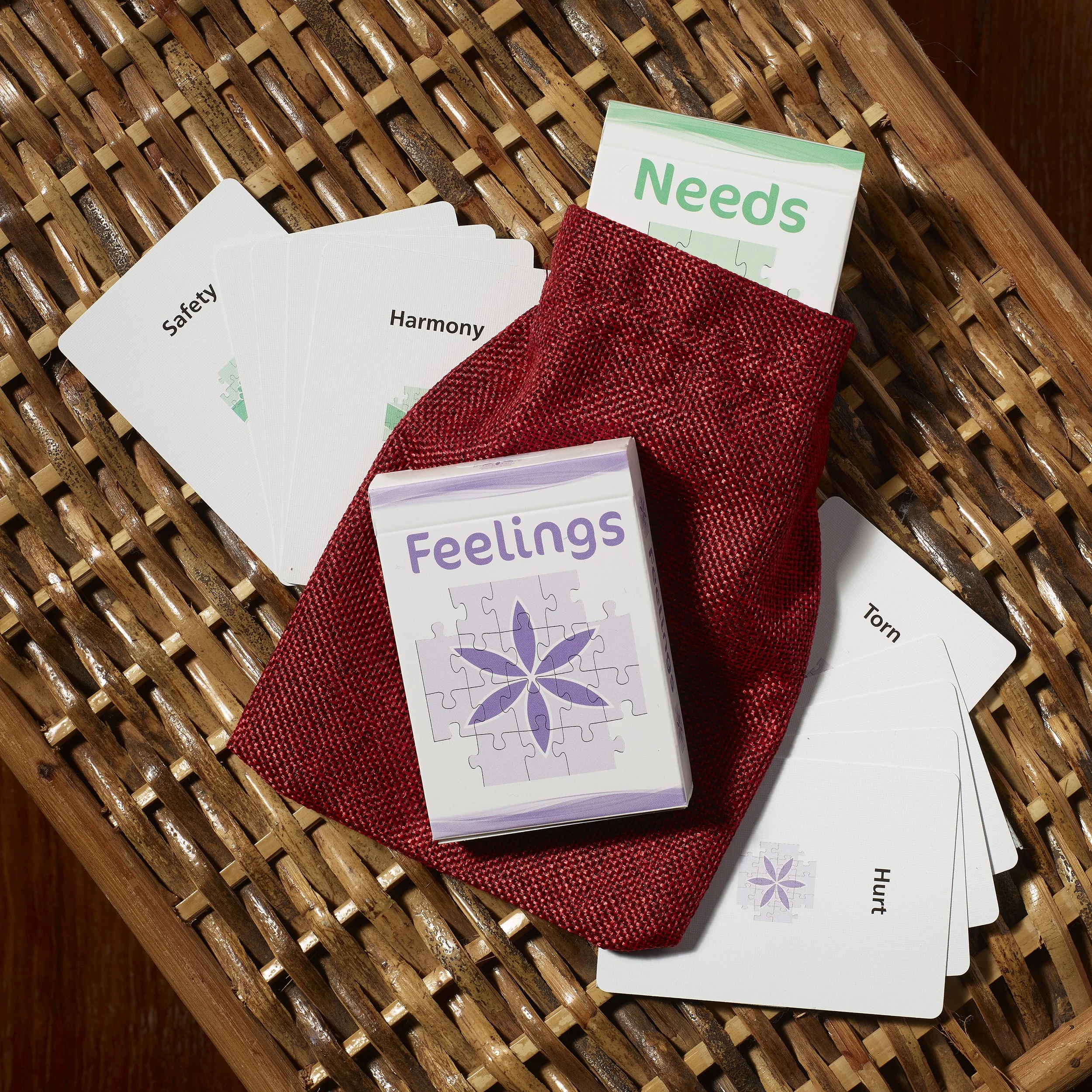 A wicker surface holding a red cloth pouch filled with card game boxes and cards. The cards include words like "Safety," "Harmony," "Needs," "Feelings," "Hurt," "Torn," and other words, suggesting an emotional or therapy-themed card game.