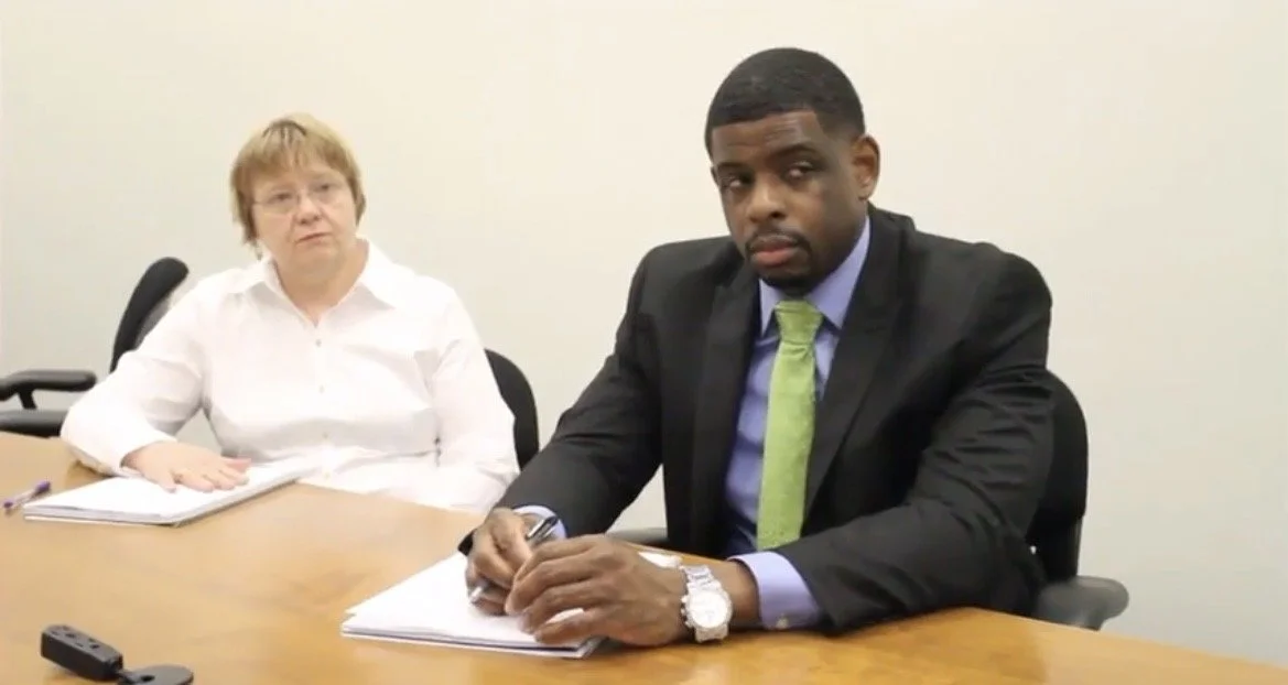 A man and a woman sitting at a conference table in an office, with the man in professional attire and the woman in a white shirt, appearing to participate in a meeting or interview.