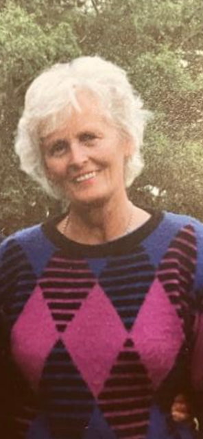 John Ford's mother, Anne, smiling with white hair outdoors, wearing a colorful striped sweater with a diamond pattern.