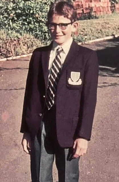John Ford with glasses wearing a school uniform (Kloof High School), standing outdoors on a paved area with greenery and a brick wall in the background.