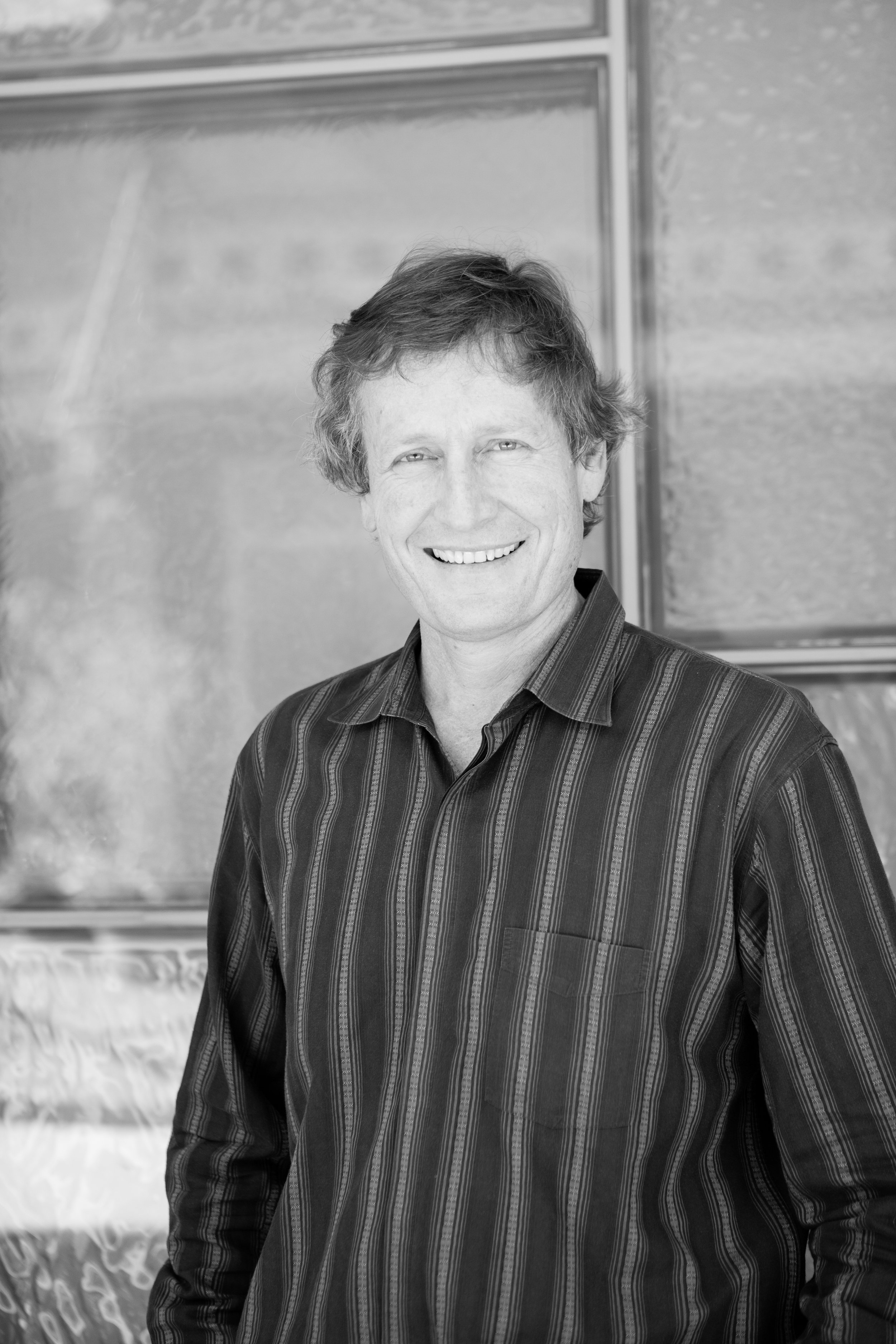 Black and white portrait of a smiling man with wavy hair wearing a striped button-up shirt, standing in front of a chalkboard.