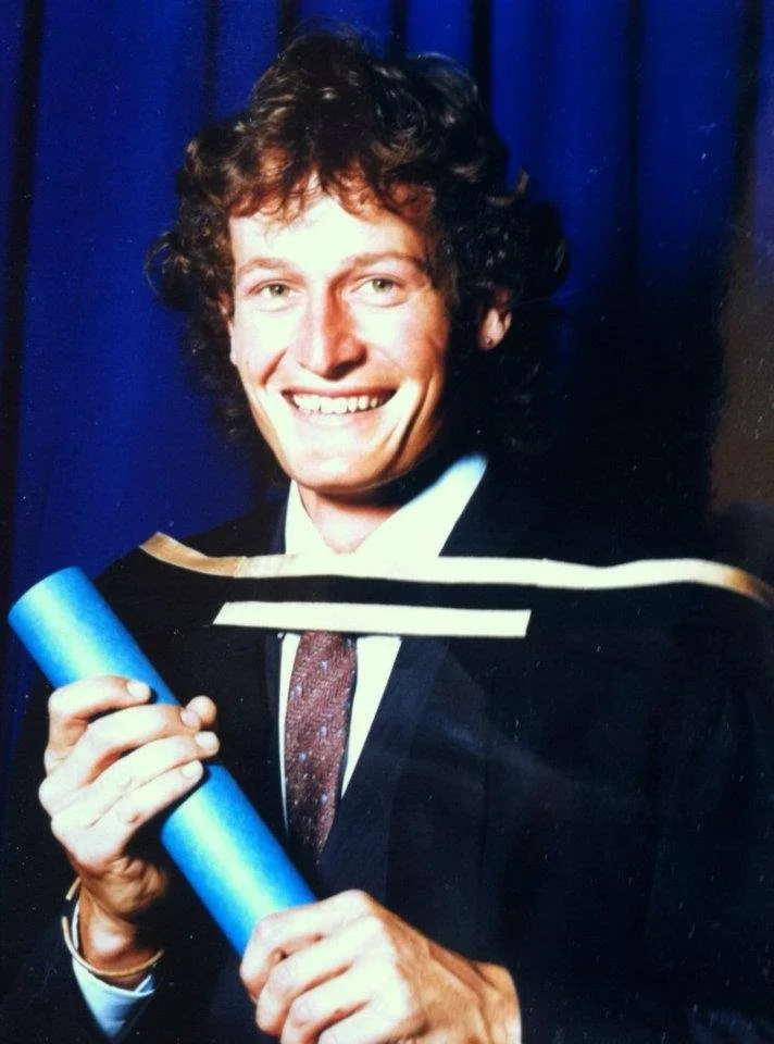 John Ford in a graduation gown holding his Law degree, smiling, with a dark blue curtain background.