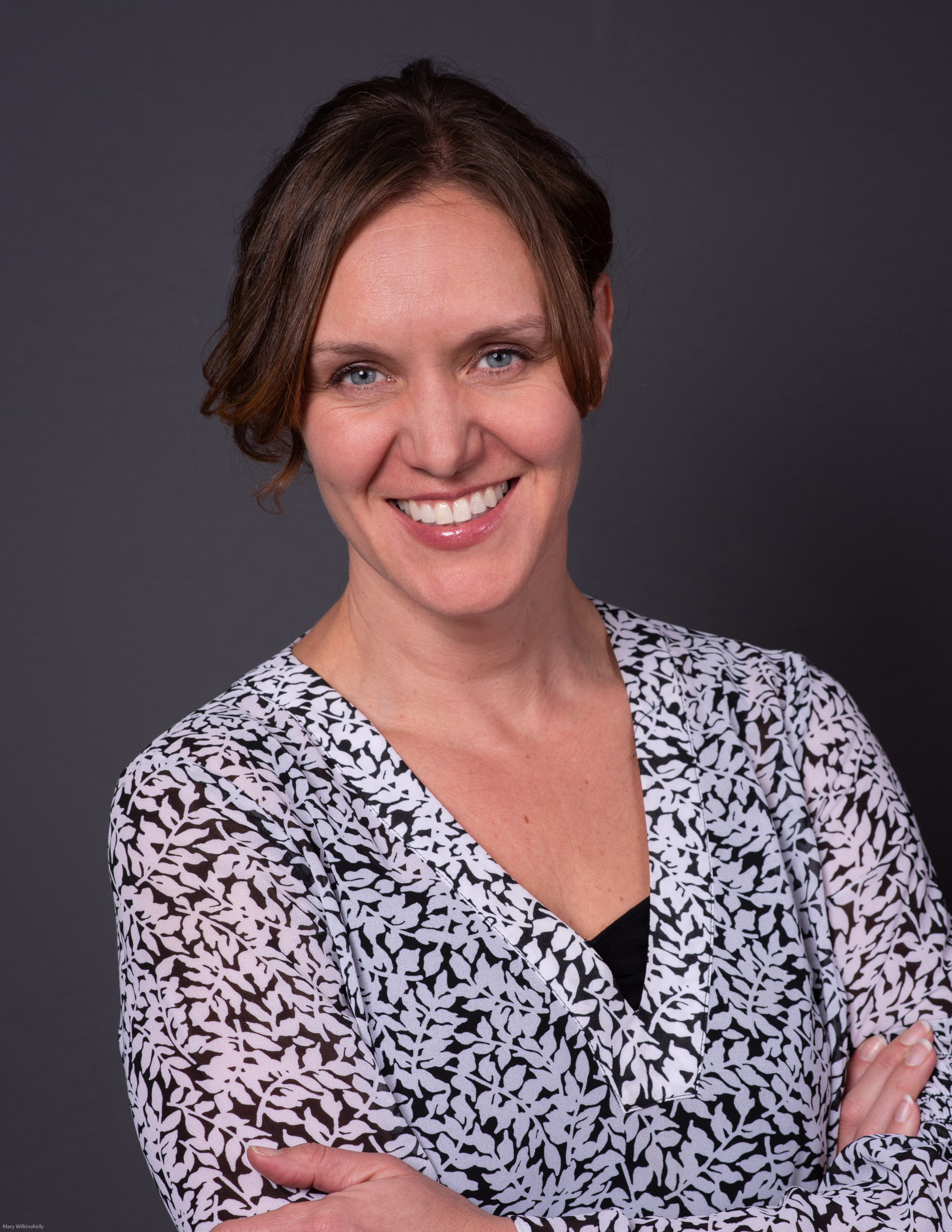 A woman with short brown hair smiling with crossed arms, wearing a black and white patterned top against a dark background.
