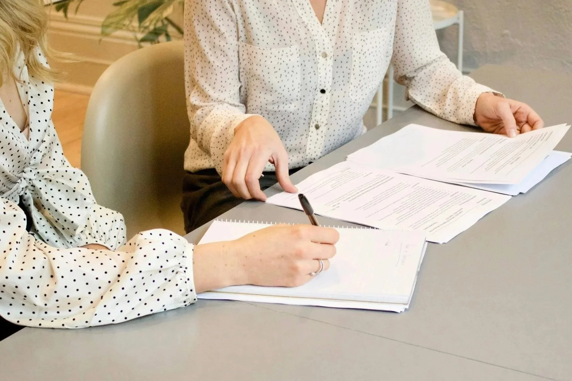 Two women sitting at a table reviewing documents and taking notes. Both women are wearing white blouses with black polka dots.