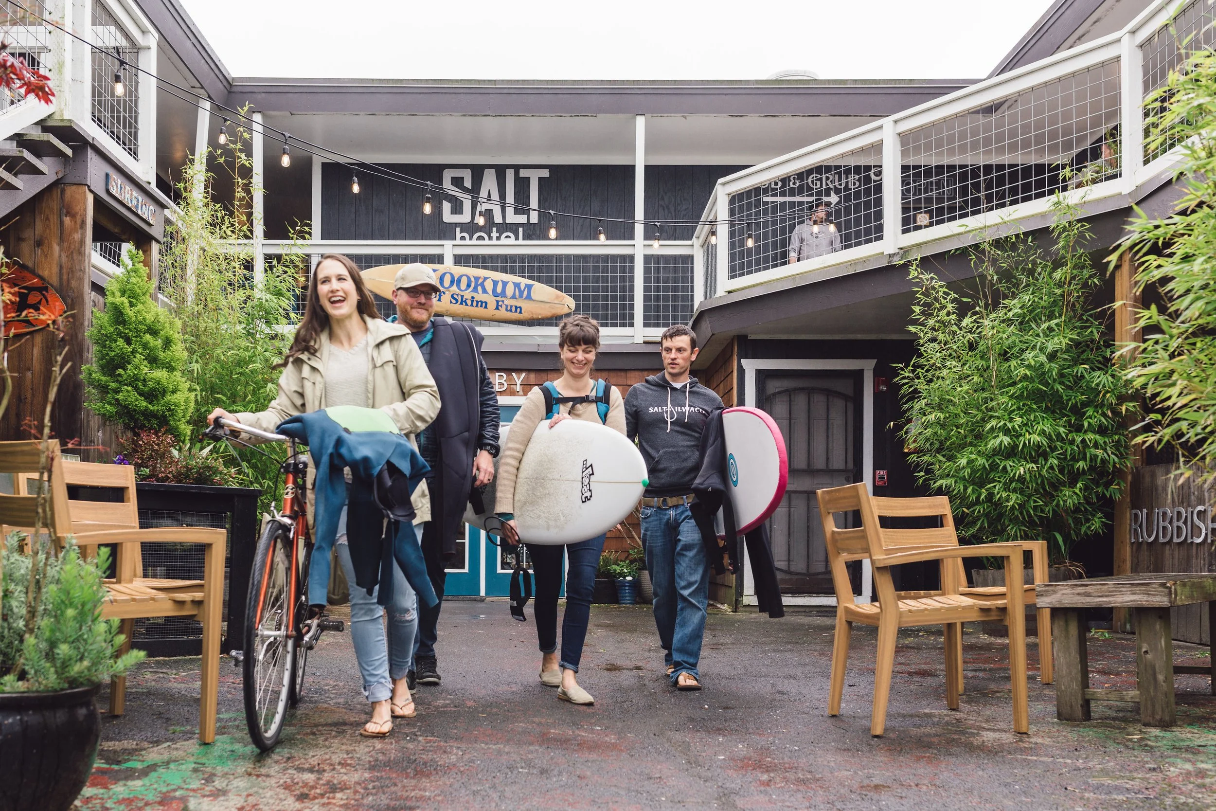 Four people, two women and two men, walking out of a surf shop, carrying surfboards and bike, on a wet outdoor patio area with chairs and plants, at a location called Salt Hotel with a salt and crab sign.