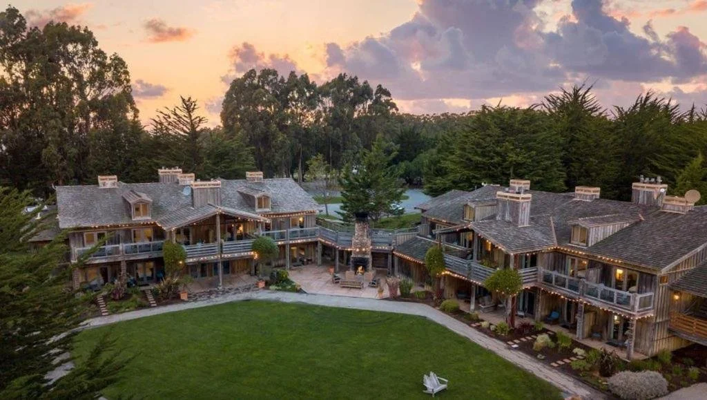 A large multi-unit rustic style house or lodge at sunset surrounded by trees, with a spacious green lawn, outdoor seating, and a central outdoor fireplace.
