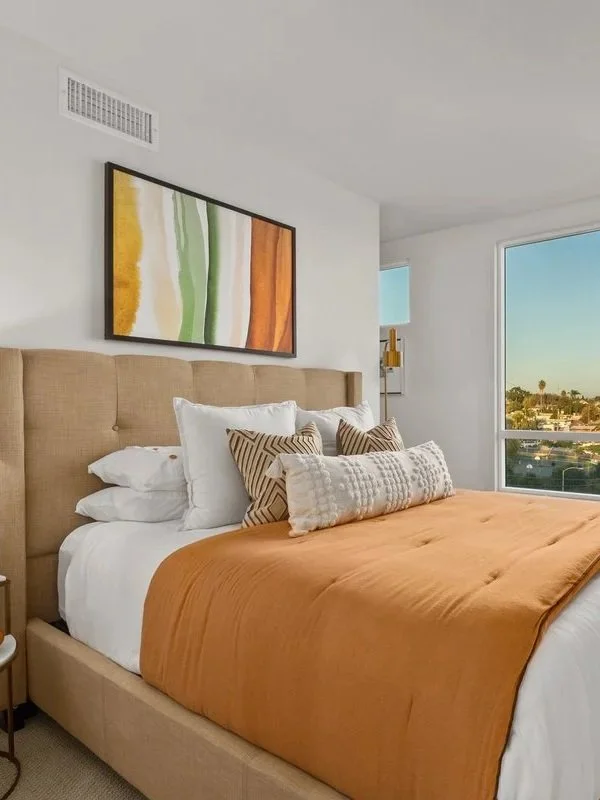 A modern bedroom featuring a beige upholstered bed with white and patterned pillows, an orange bedspread, a nightstand with a white lamp and decorative items, a large window showing a cityscape view, and a framed abstract artwork on the wall.