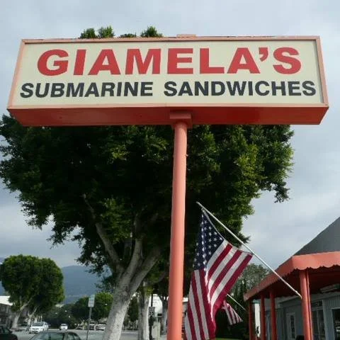 Sign for Giamela's Submarine Sandwiches with an American flag hanging nearby, outside a restaurant with trees and a cloudy sky in the background.