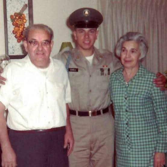 Three people posing together indoors: an elderly man on the left, a young man in the middle in military uniform, and an elderly woman on the right. The background features curtains and decorative wall hangings.