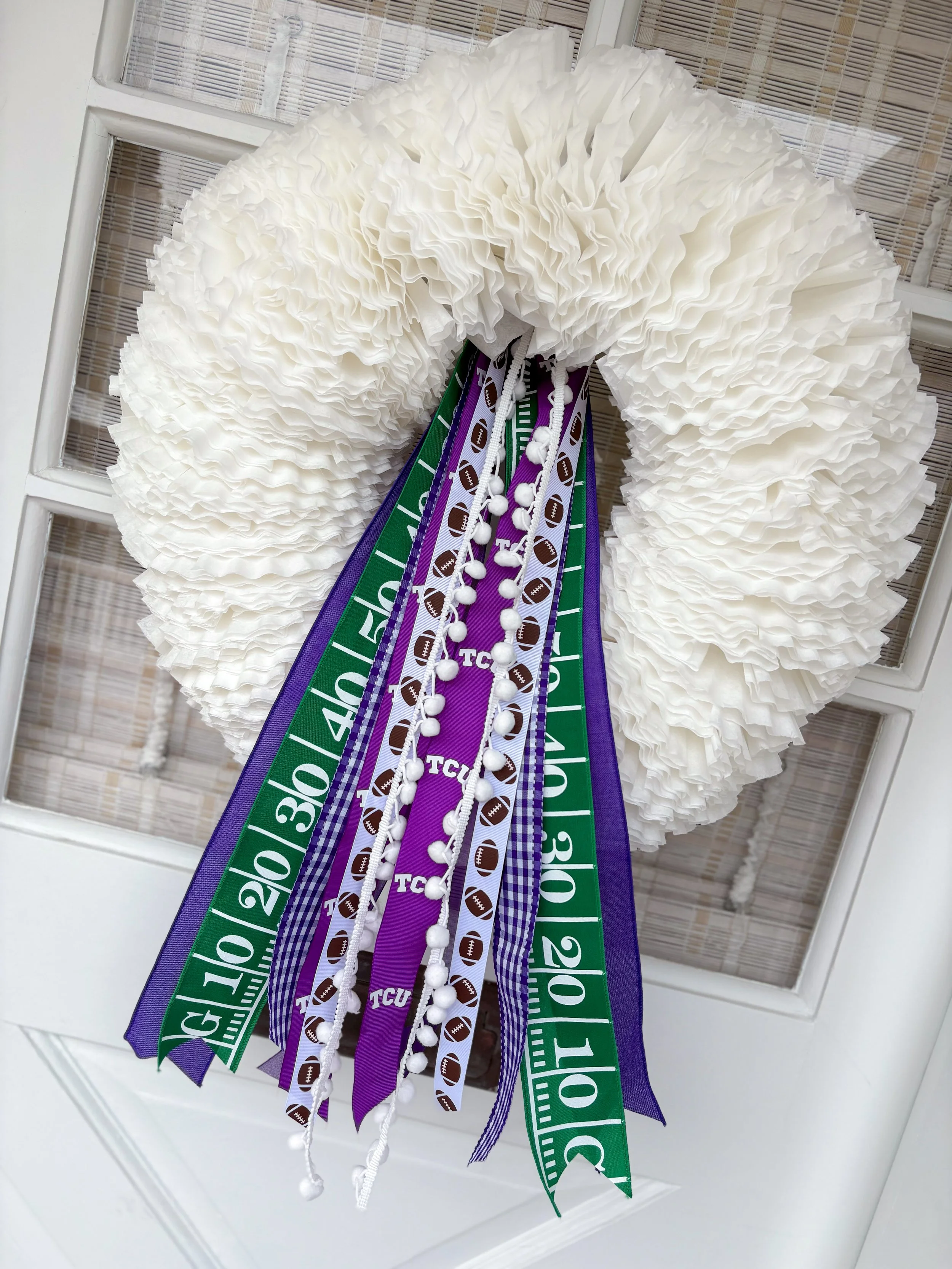 White decorative wreath with purple, green, and purple-and-white ribbons with football graphics and 'TCU' text hanging, displayed on a door.