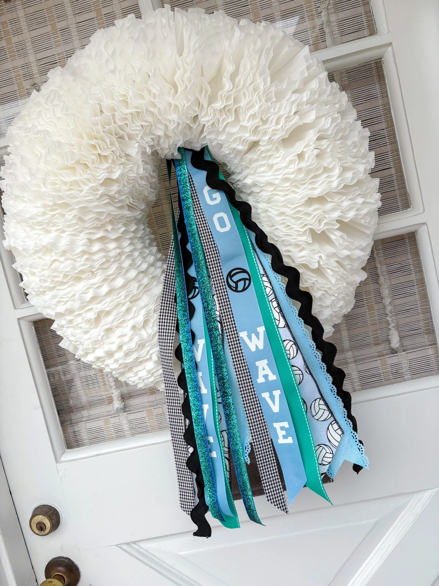 A white ruffled decorative wreath hangs on a glass-paneled white door, adorned with blue, black, and patterned ribbons with words wave and volleyball illustrations.