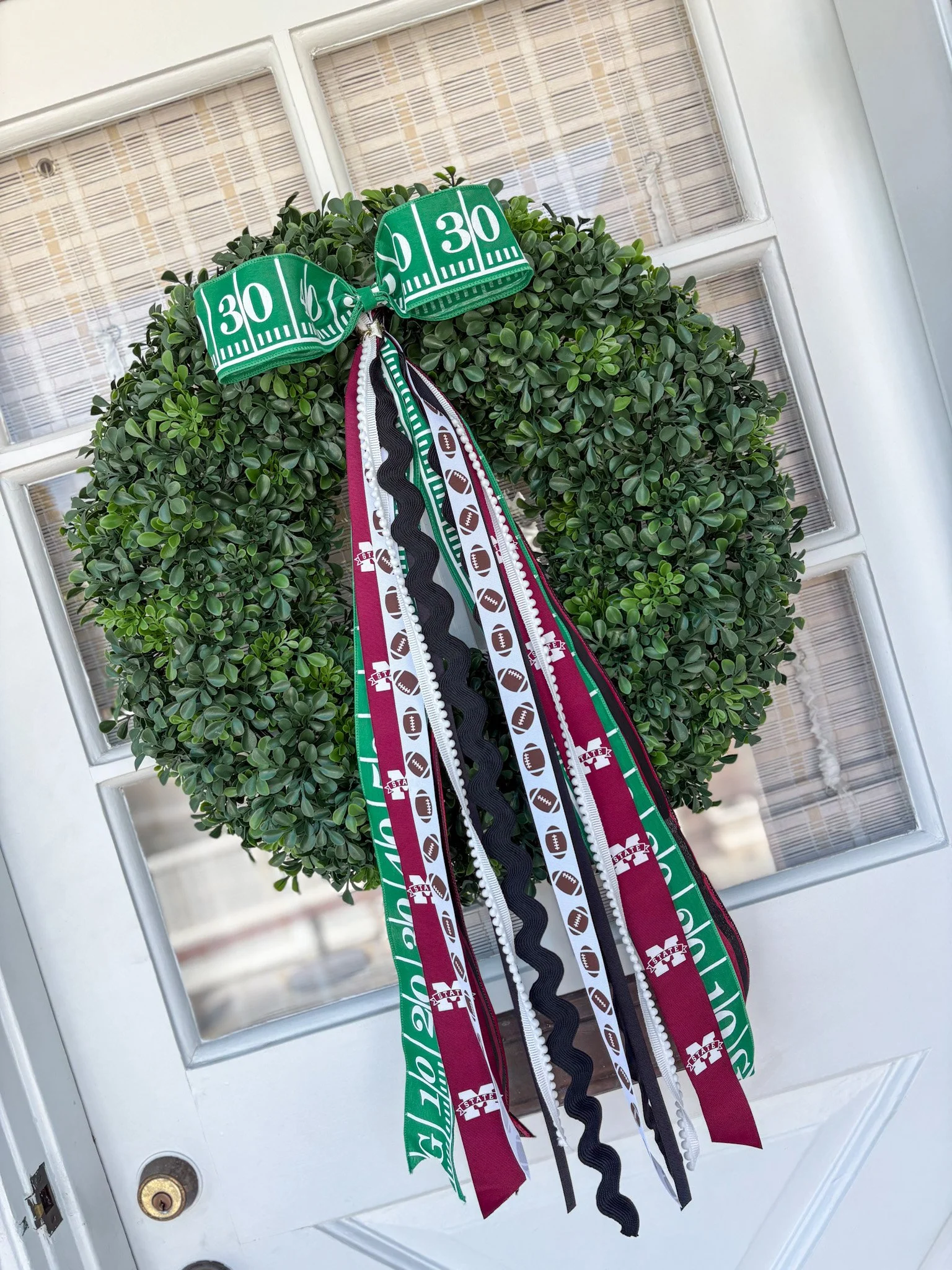 Green decorative wreath with ribbons and fabric strips featuring football and football team logos, hanging on a door.