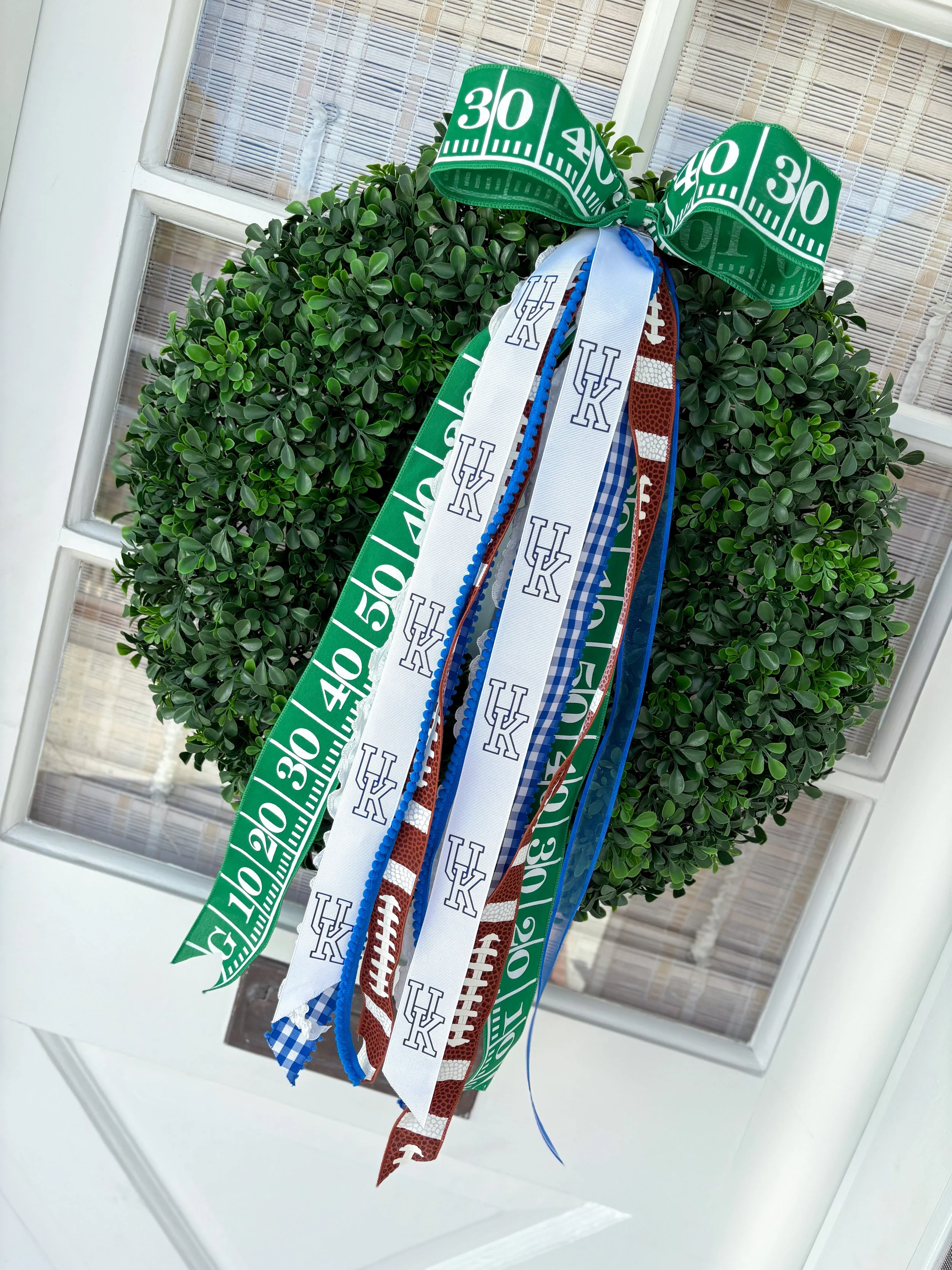 Decorative wreath with green, white, and blue ribbons, including a green ribbon with white numbers, a white ribbon with blue 'UK' logos, and a brown football-patterned ribbon, hanging on a white door with a glass panel.