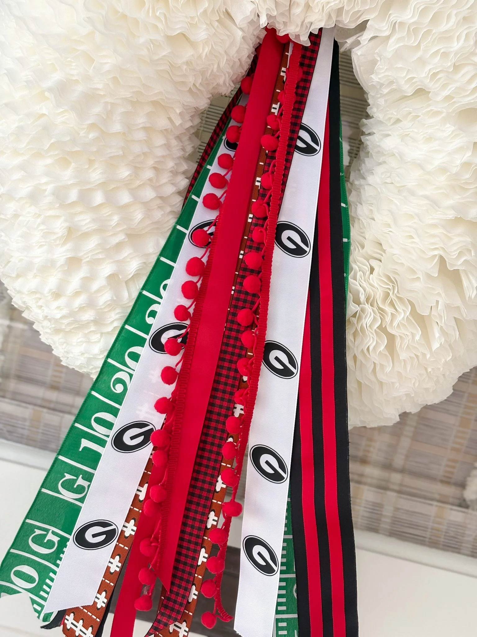 Various patterned and colored ribbons hanging, including green, white, black, red, and checkered designs, with some ribbons featuring pom-pom details, in front of a white, curly textured background.
