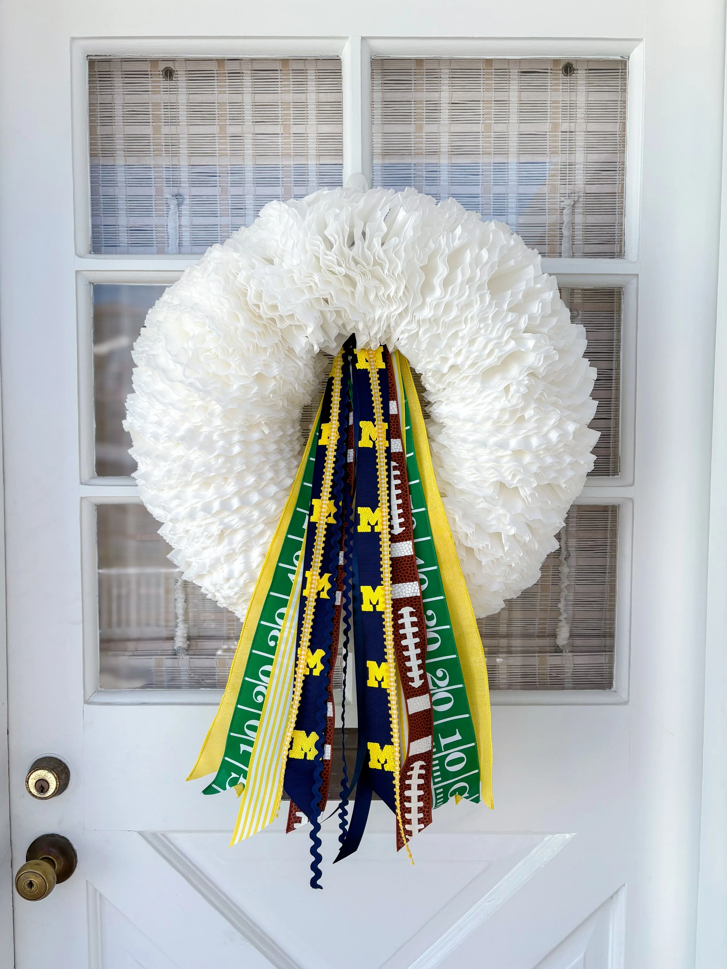 A white wreath decorated with University of Michigan-themed ribbons, hanging on a door.