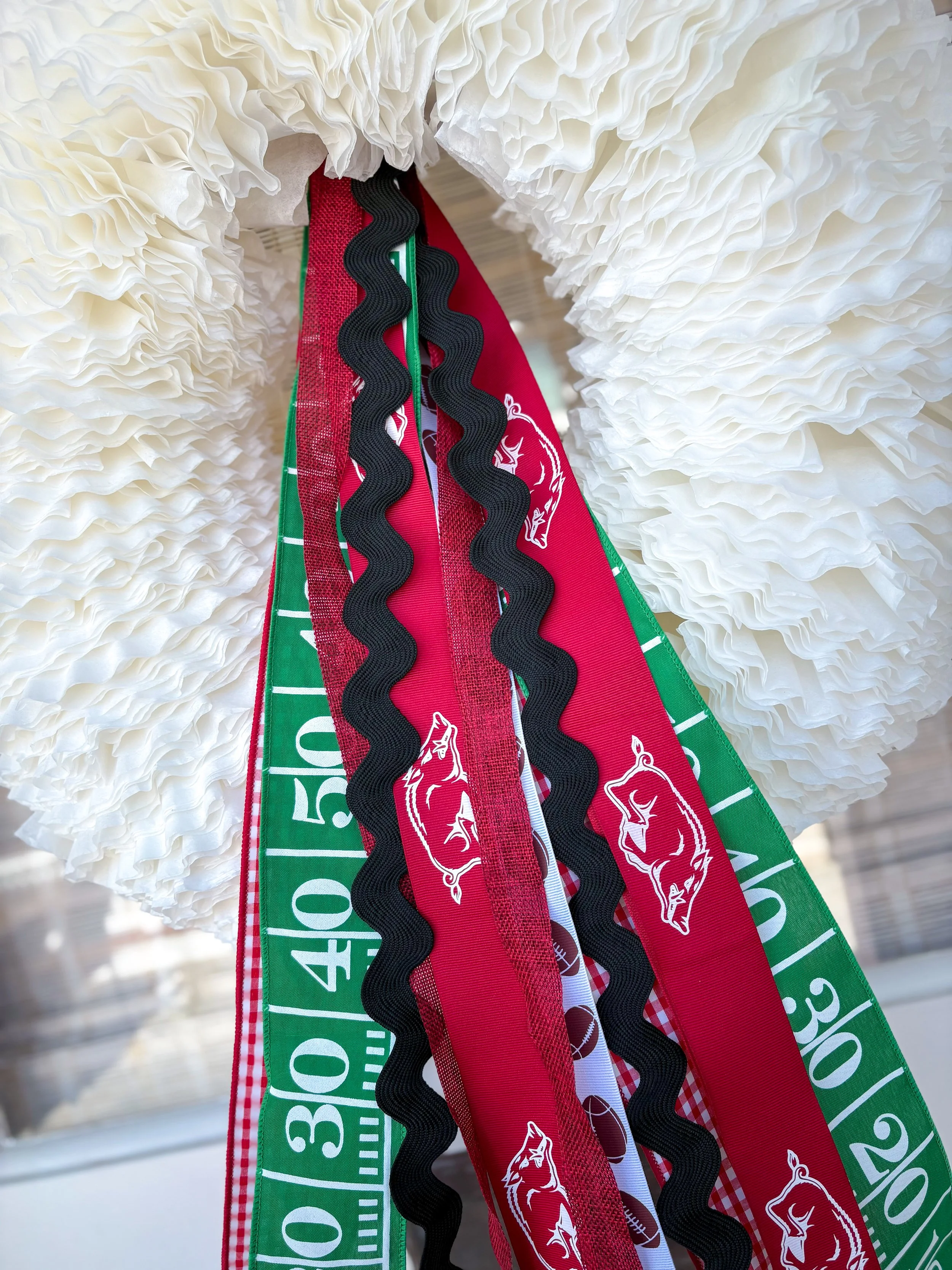 Close-up of various decorative ribbons hanging from a white textured paper pom-pom ornament, with the ribbons featuring football-themed and Arkansas.