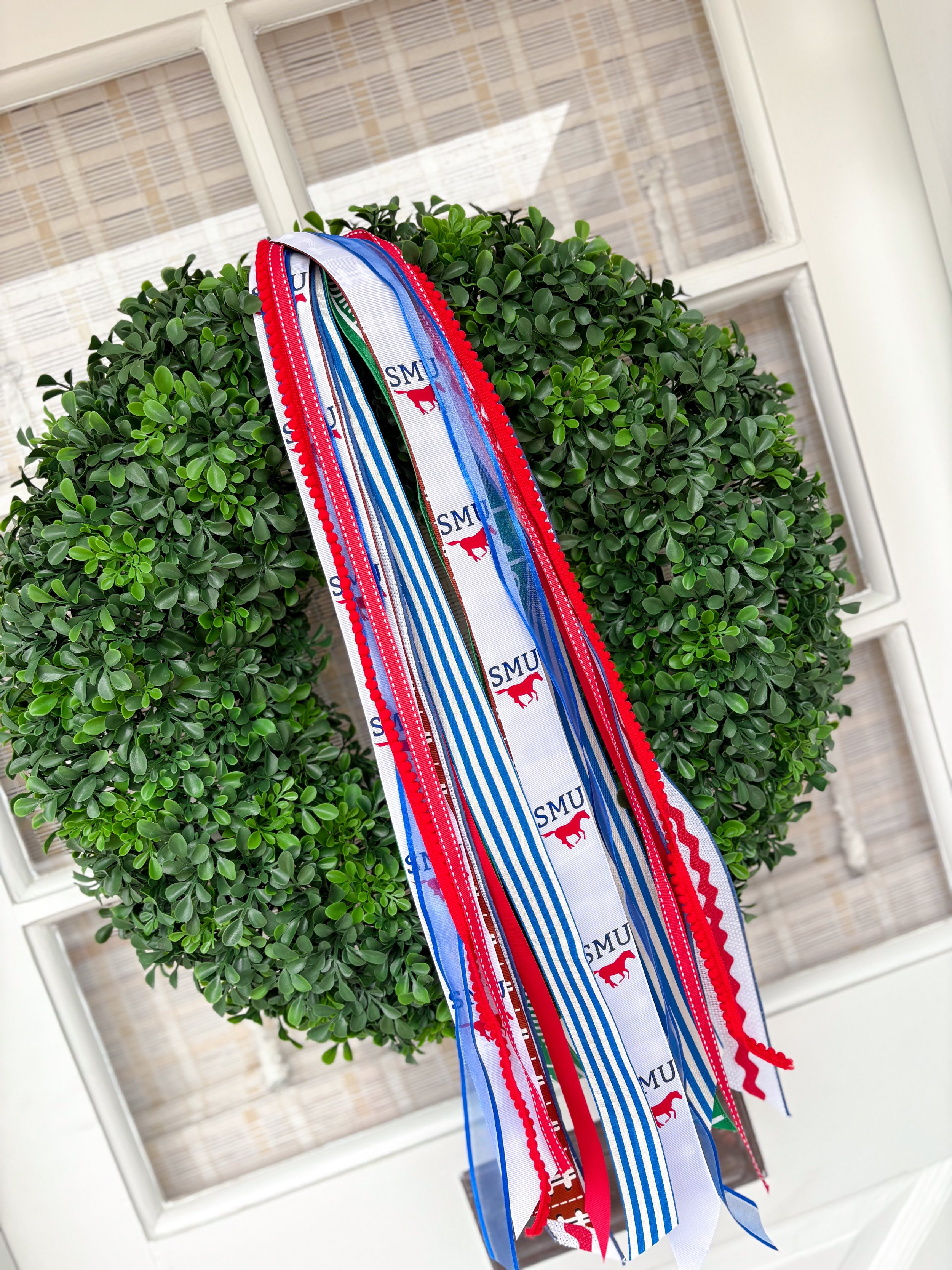 Green leafy topiary shrub with multiple colorful ribbons hanging from it, some with blue and white stripes, others with red edges and the acronym "SMU" and a red horse logo, in front of a white window with blinds.