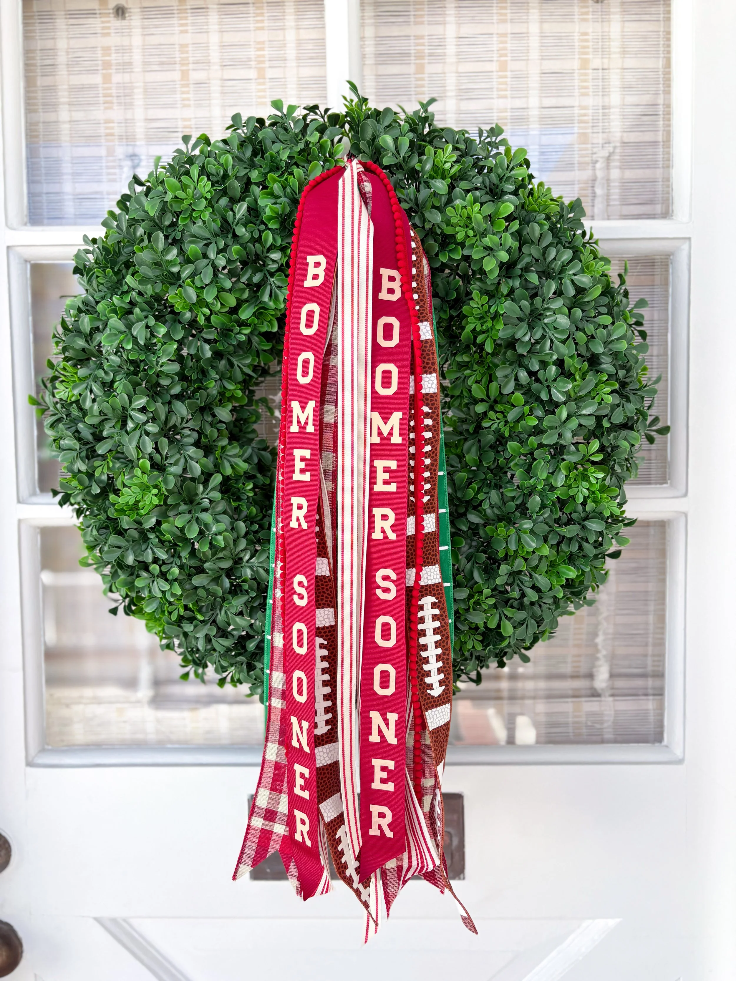 Decorative wreath with red and white ribbons that have "Boomer Sooner" written on them, hanging on a white door with glass panels.