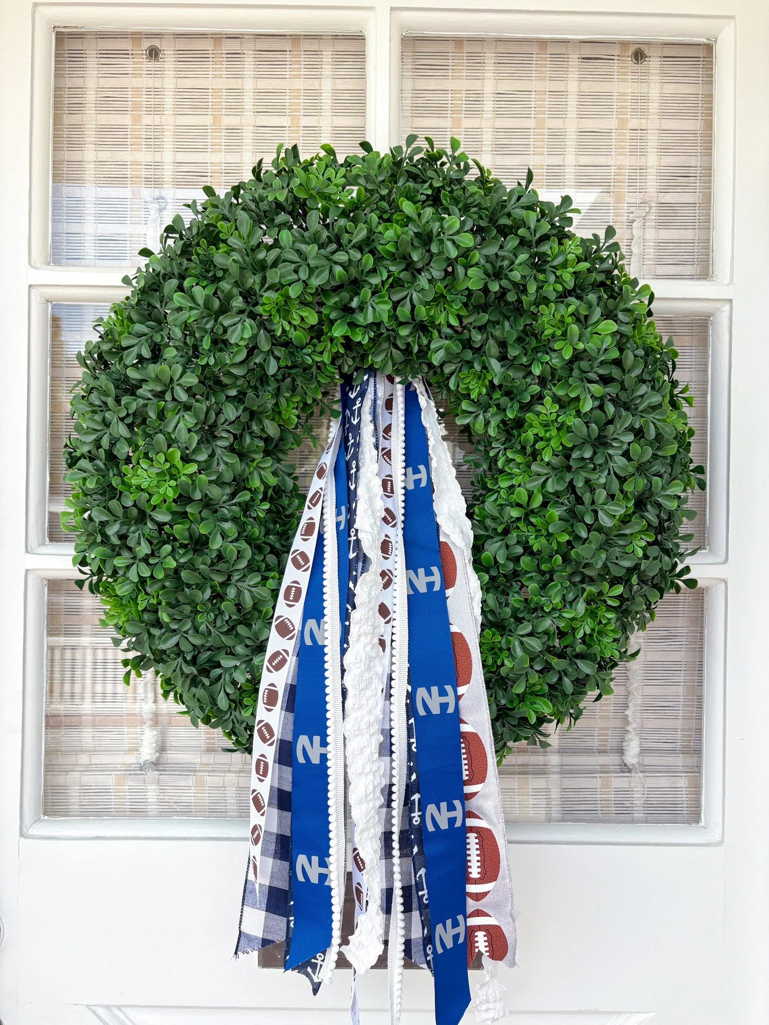 Green decorative wreath with hanging fabric ribbons featuring Newport harbor football and team logos, hung on a white door with glass panels and bamboo blinds.