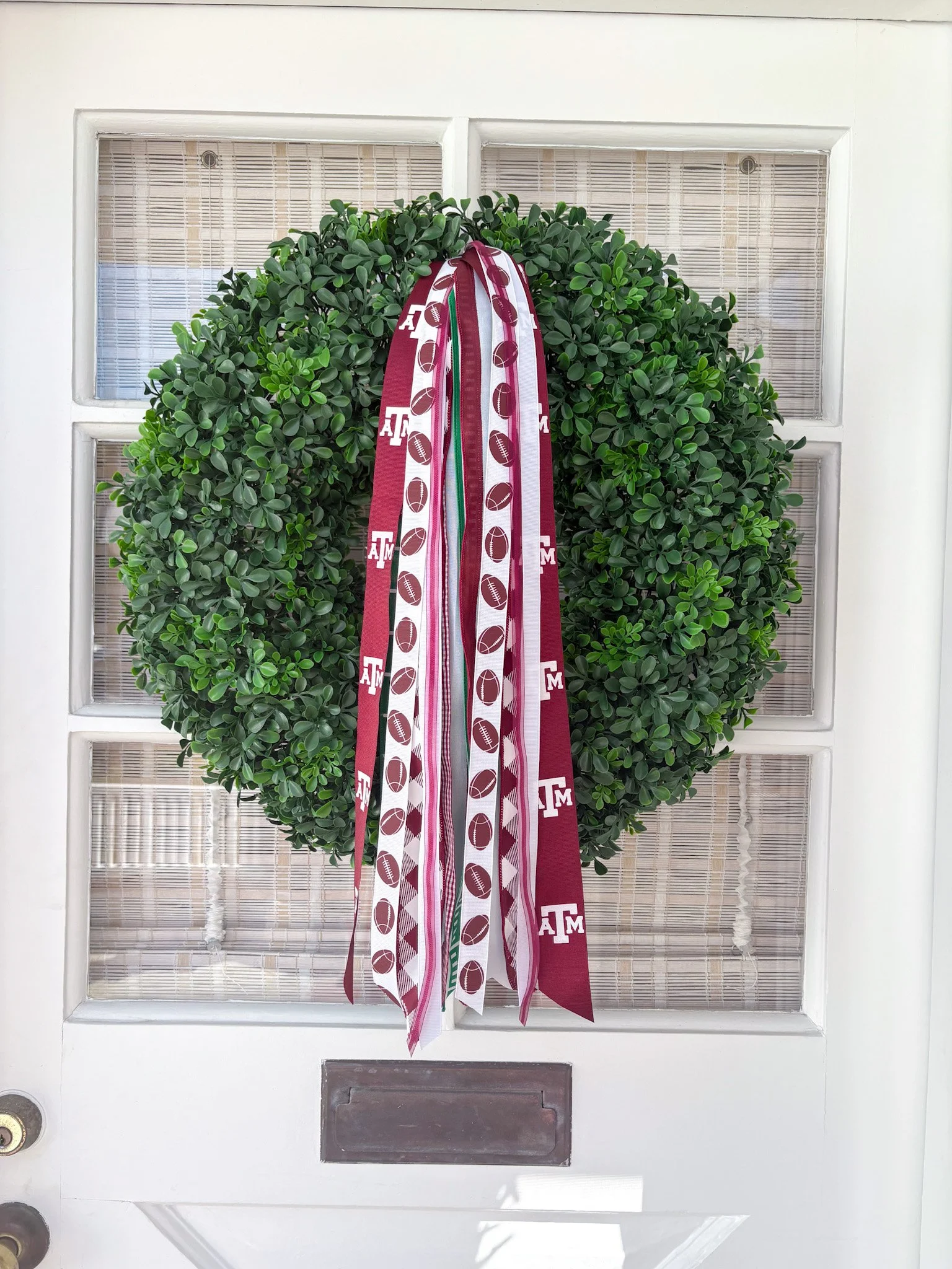 Green boxwood wreath decorated with ribbons featuring A&M University football and logo, hung on a white door.