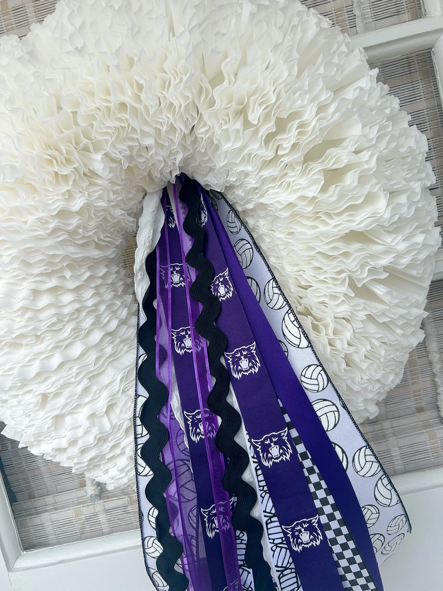 A decorative wreath made of white ruffled fabric with purple, black, and white volleyball ribbons hanging from the center, some featuring a sports team's wildcat logo.