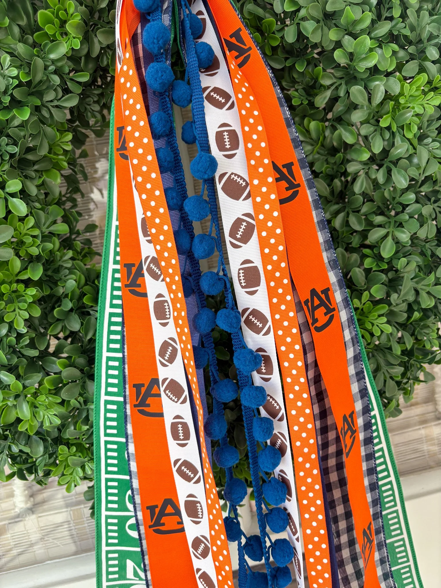Close-up of decorative ribbons with sports themes, including footballs, University, and patterns in blue, orange, white, and green colors, hanging on a faux green wreath.