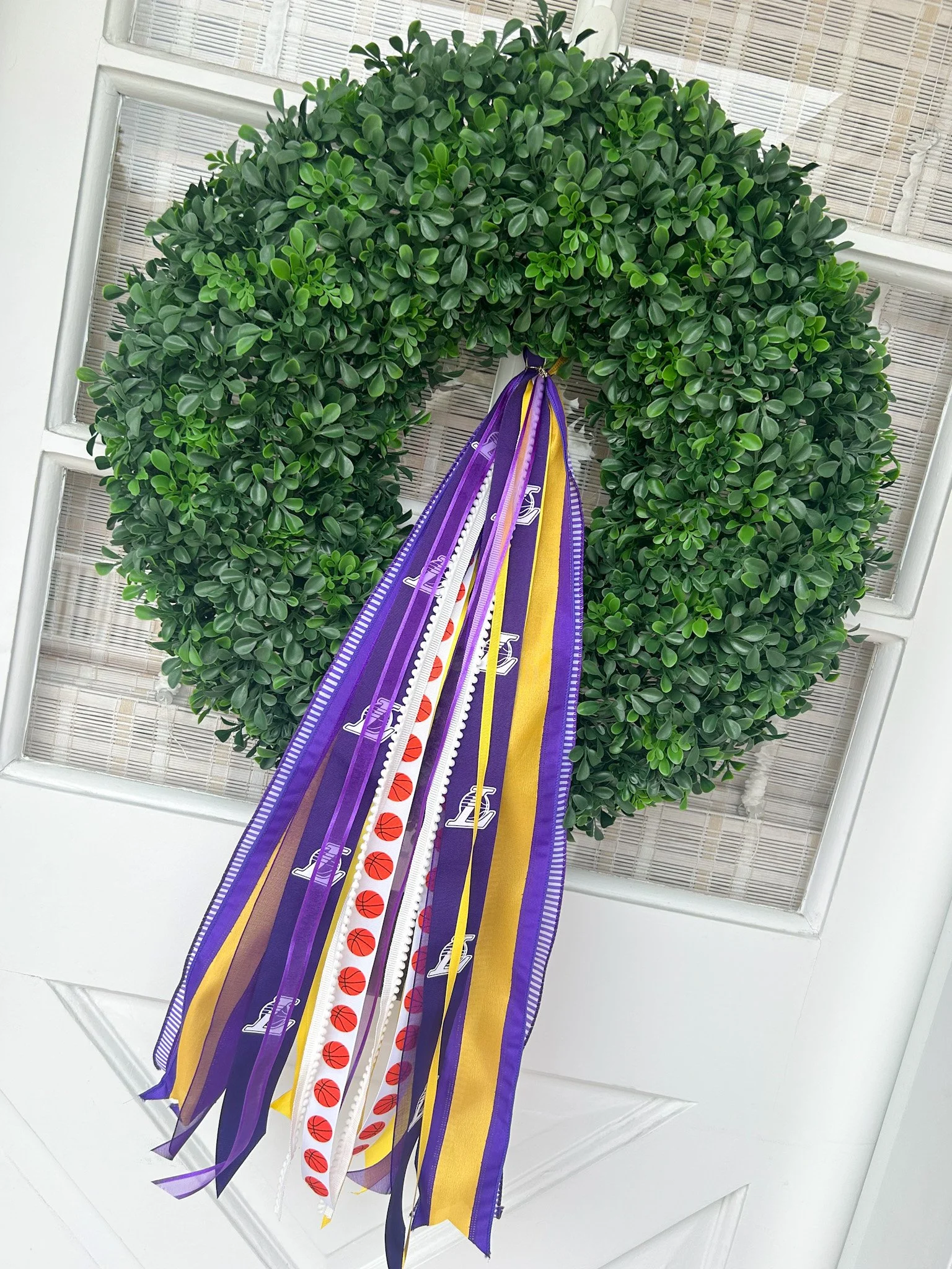 Green greenery wreath with purple, yellow, white, and basketball-themed ribbons hanging on a white door.