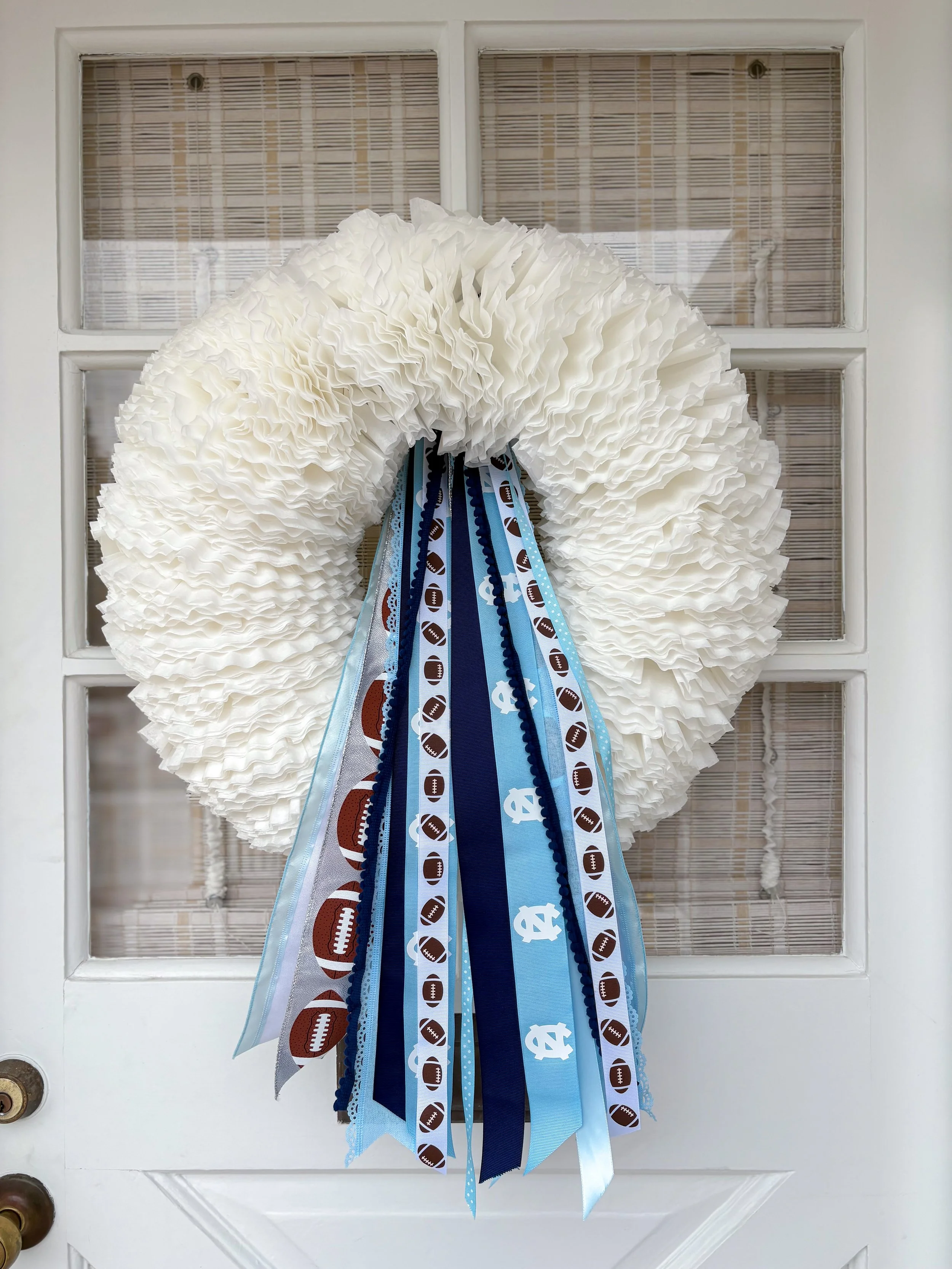 A white, ruffled tissue paper wreath decorated with light blue and dark blue ribbon strips, including football patterns and NC  Heels symbols, hung on a white door.