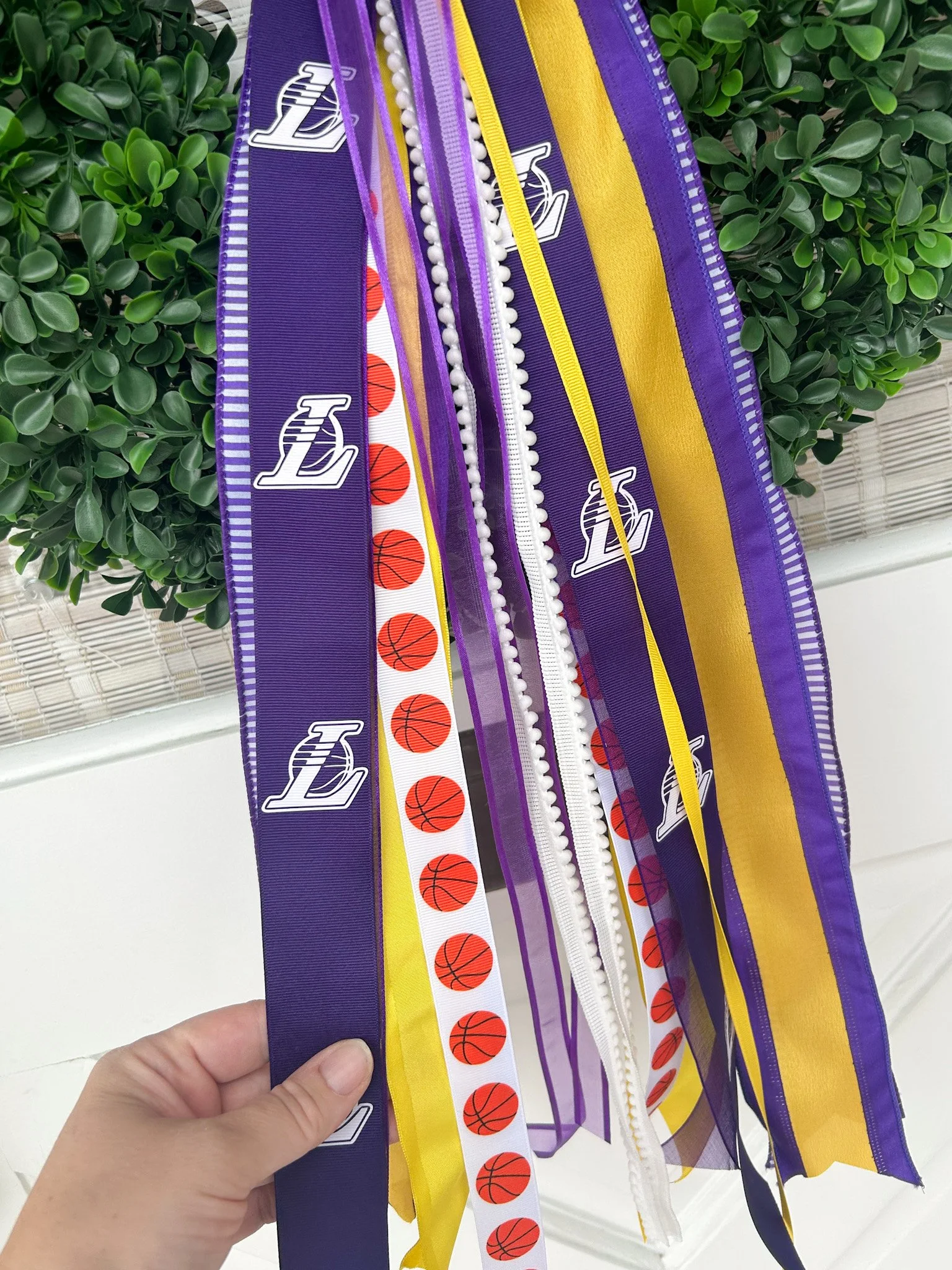 Hand holding a collection of colorful Los Angeles basketball-themed ribbons, including purple, yellow, white with basketball prints, and some with logo.