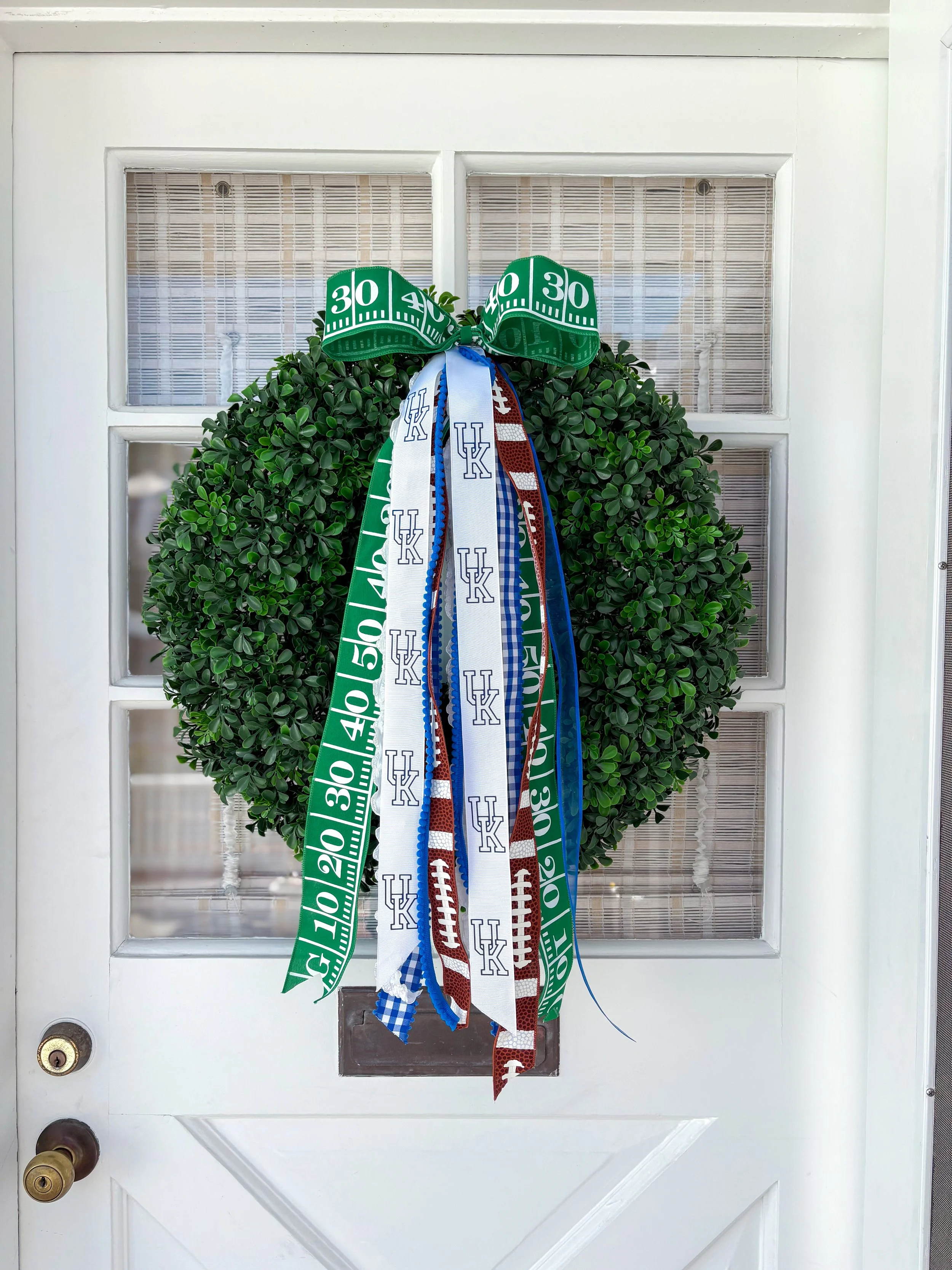 A door decorated with a green wreath and a large bow made of green and white ribbon with football field markings, along with various blue, white, and red ribbons featuring a Kentucky and football motifs.