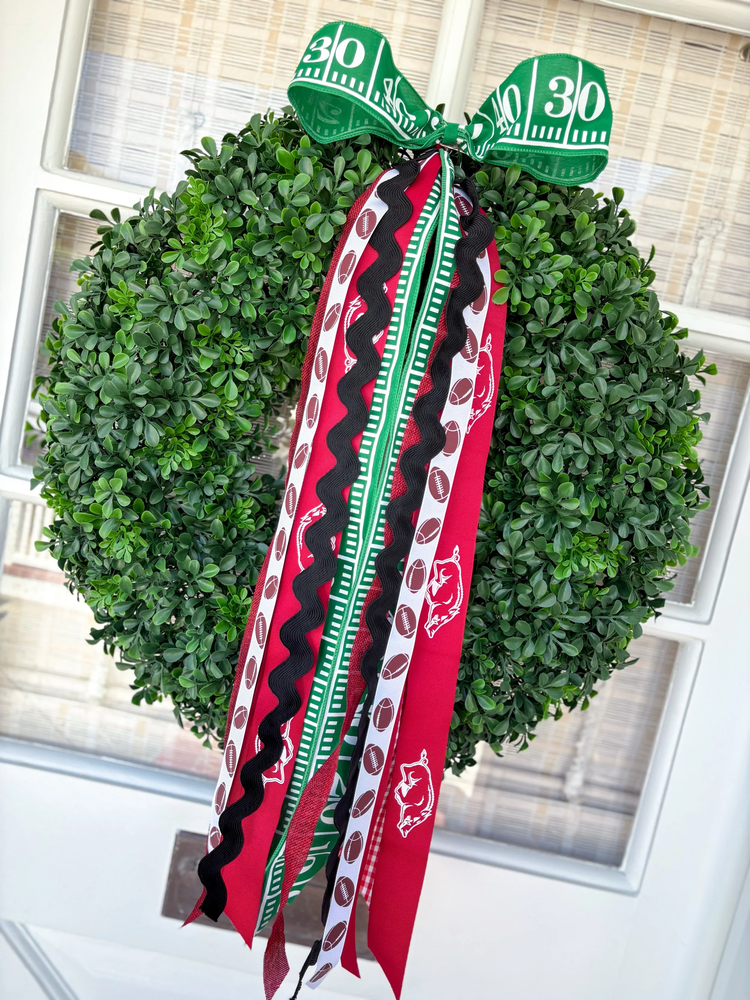 Faux green boxwood wreath decorated with ribbons in red, green, black, and white, featuring football graphics and Arkansas, hanging on a door.