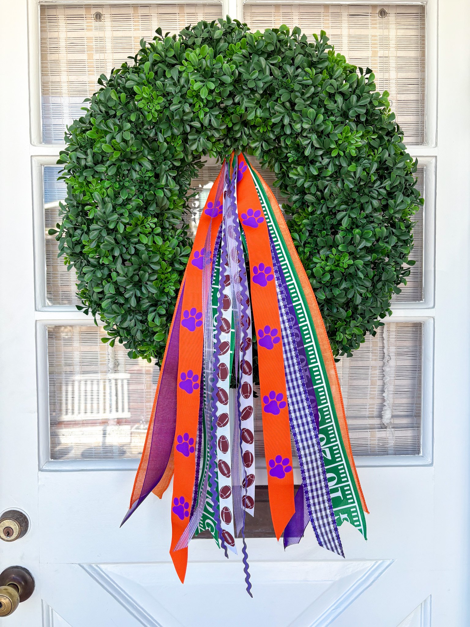 A green wreath with hanging ribbons in orange, purple, green, and purple checkered patterns, decorated with purple paw prints, attached to a white door.