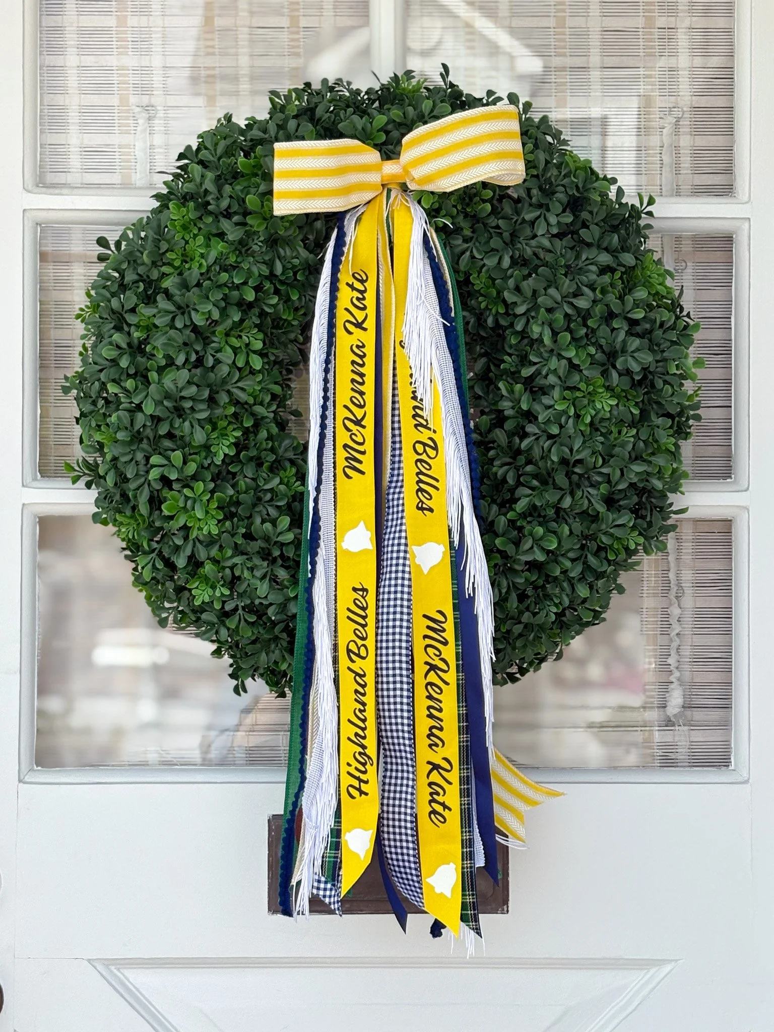 Green wreath decorated with blue, yellow, and checkered ribbons, topped with a yellow and white striped bow, featuring 'Highland belles'.