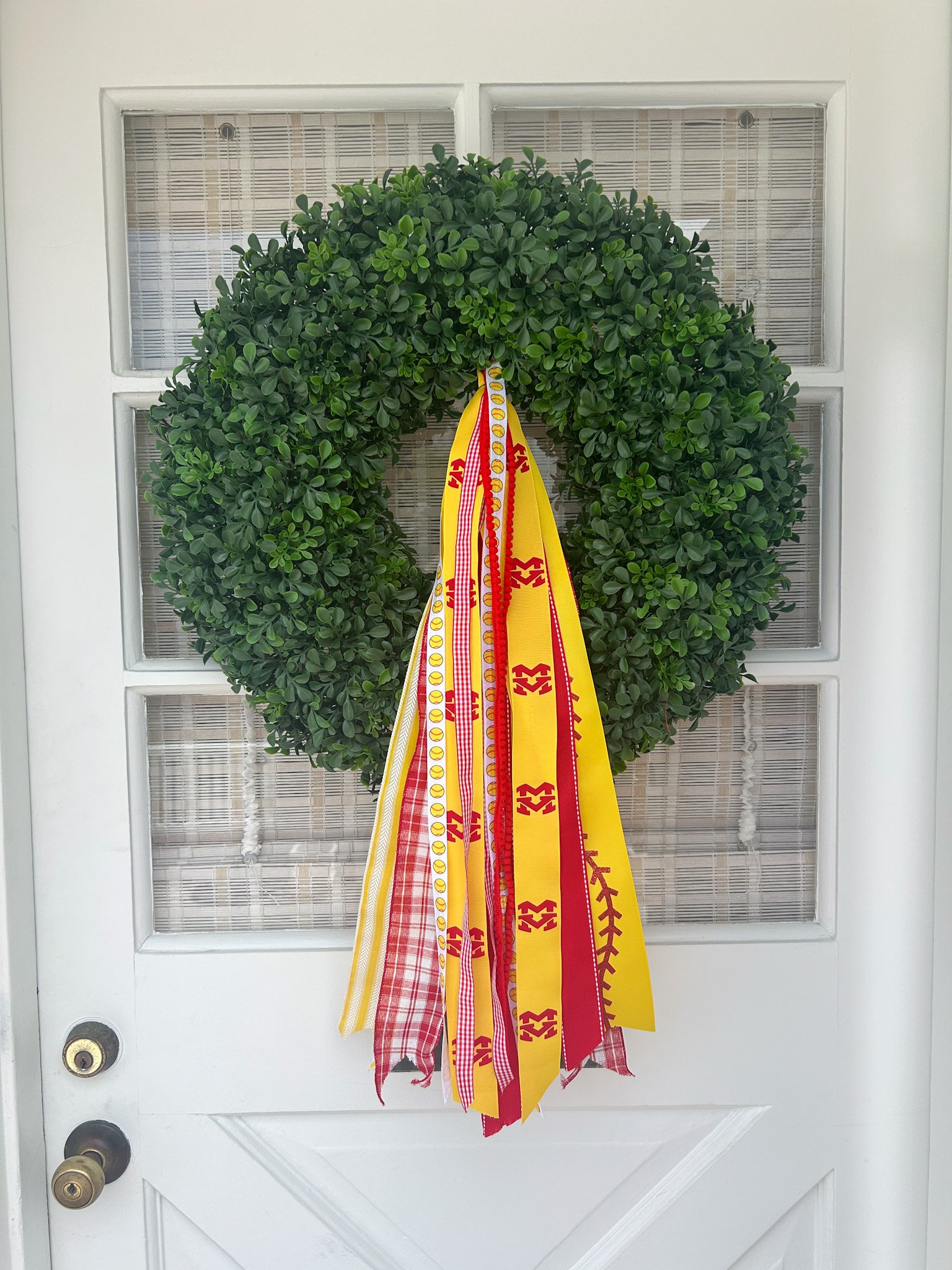 A decorative green wreath hanging on a white door with a grid window, adorned with red and yellow ribbons, including some with plaid and striped patterns.