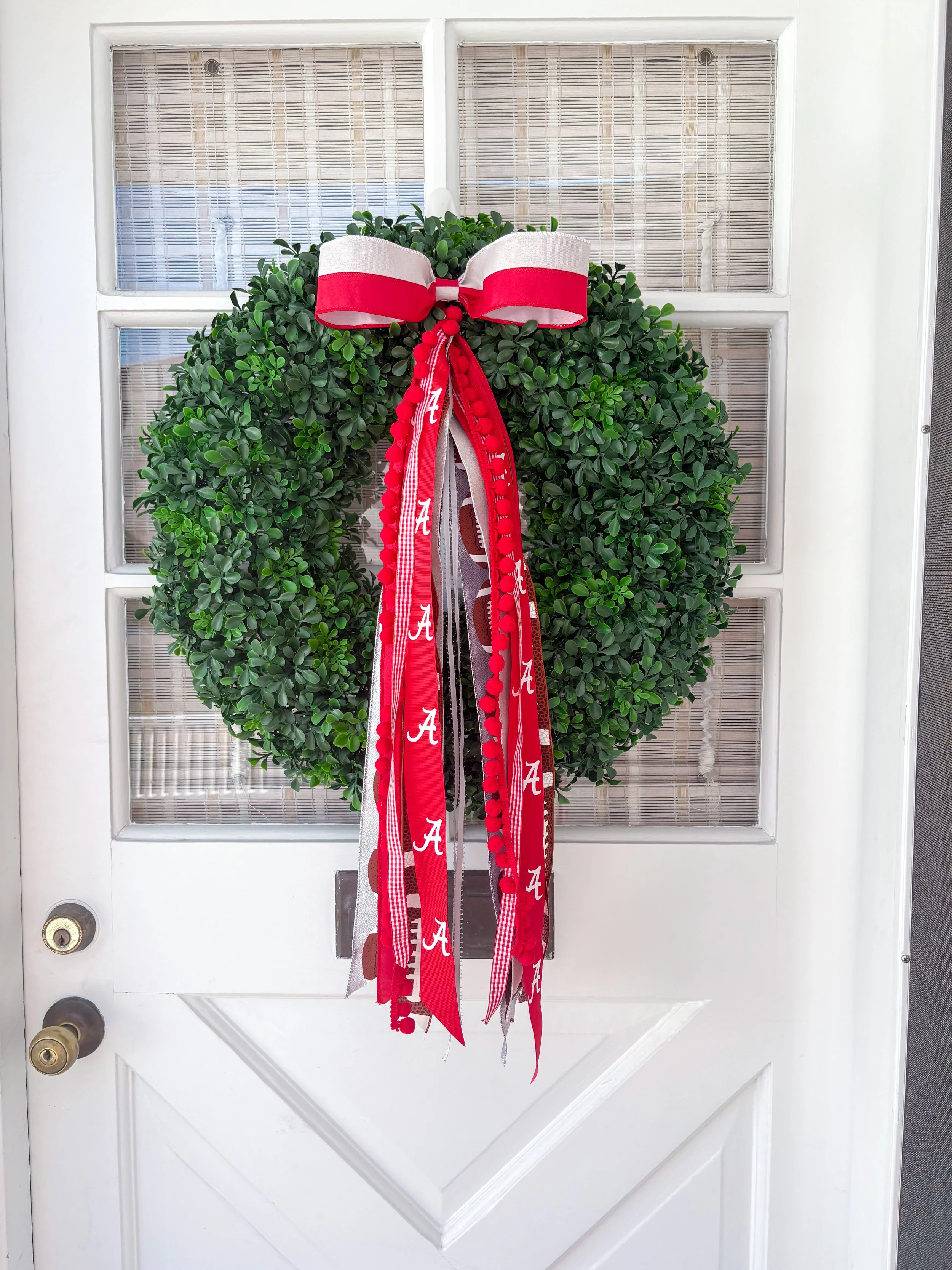 Green wreath decorated with a large white and red bow and red, white, and gray ribbons with University logos, hanging on a white door with glass panes.