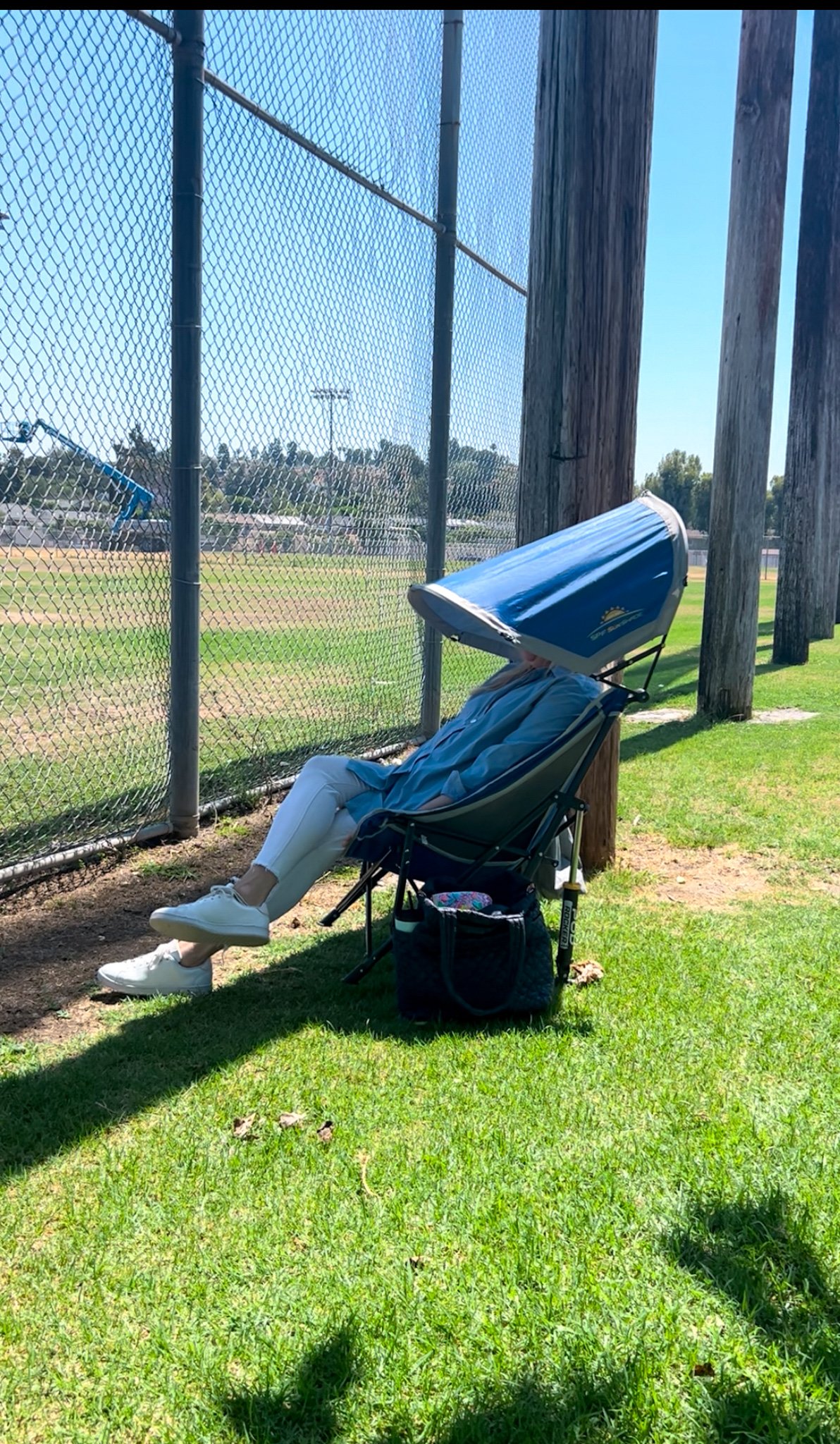 A person sitting in a chair with a large blue sunshade, watching a baseball game at a park, next to a chain-link fence and wooden poles, on a sunny day.