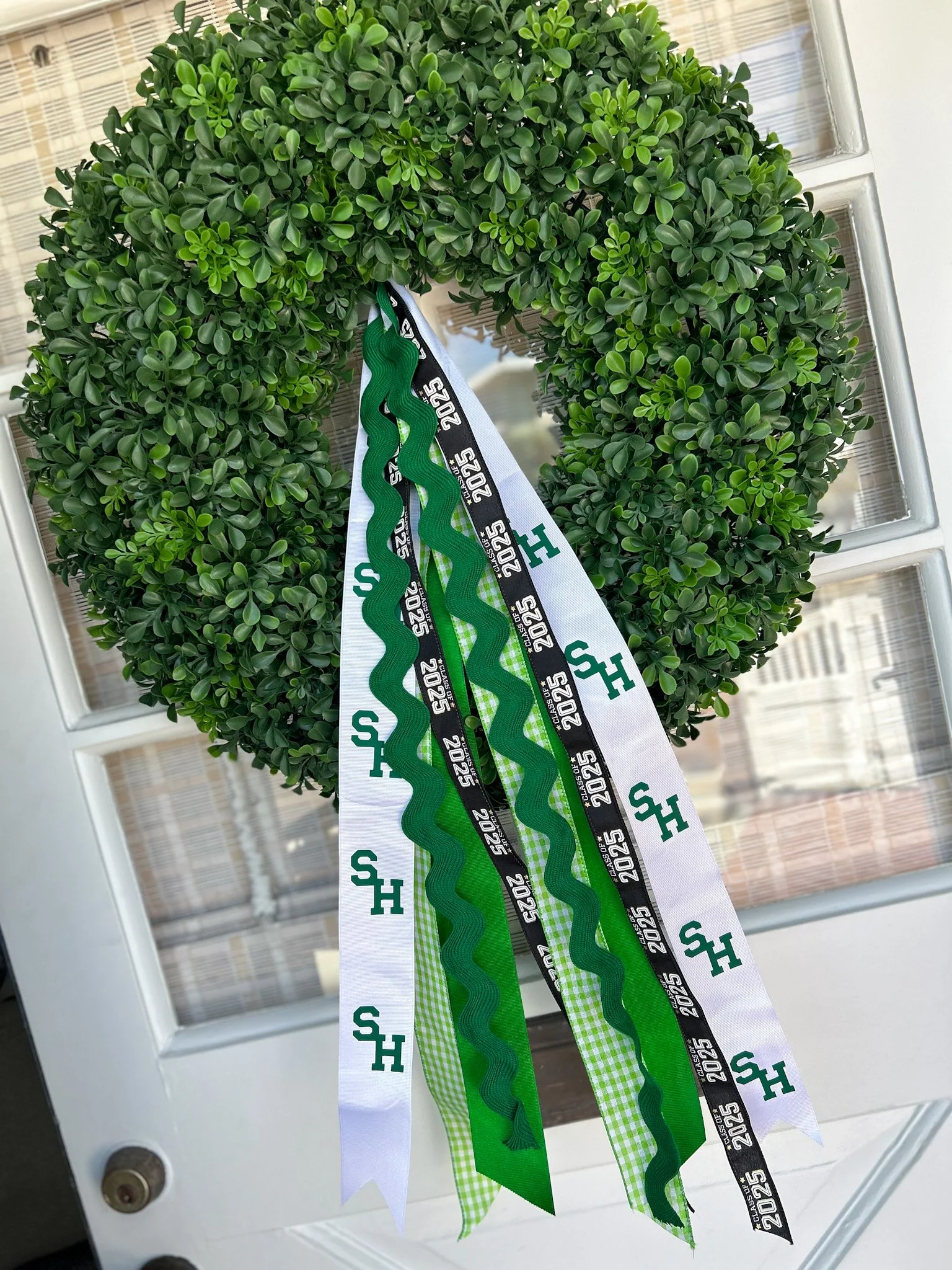 A decorative green wreath with ribbons hanging from it. The ribbons include a white ribbon with green Sage Hill logos, a black ribbon with white '2025' and other text, a dark green curly ribbon, and a checkered green and white ribbon.