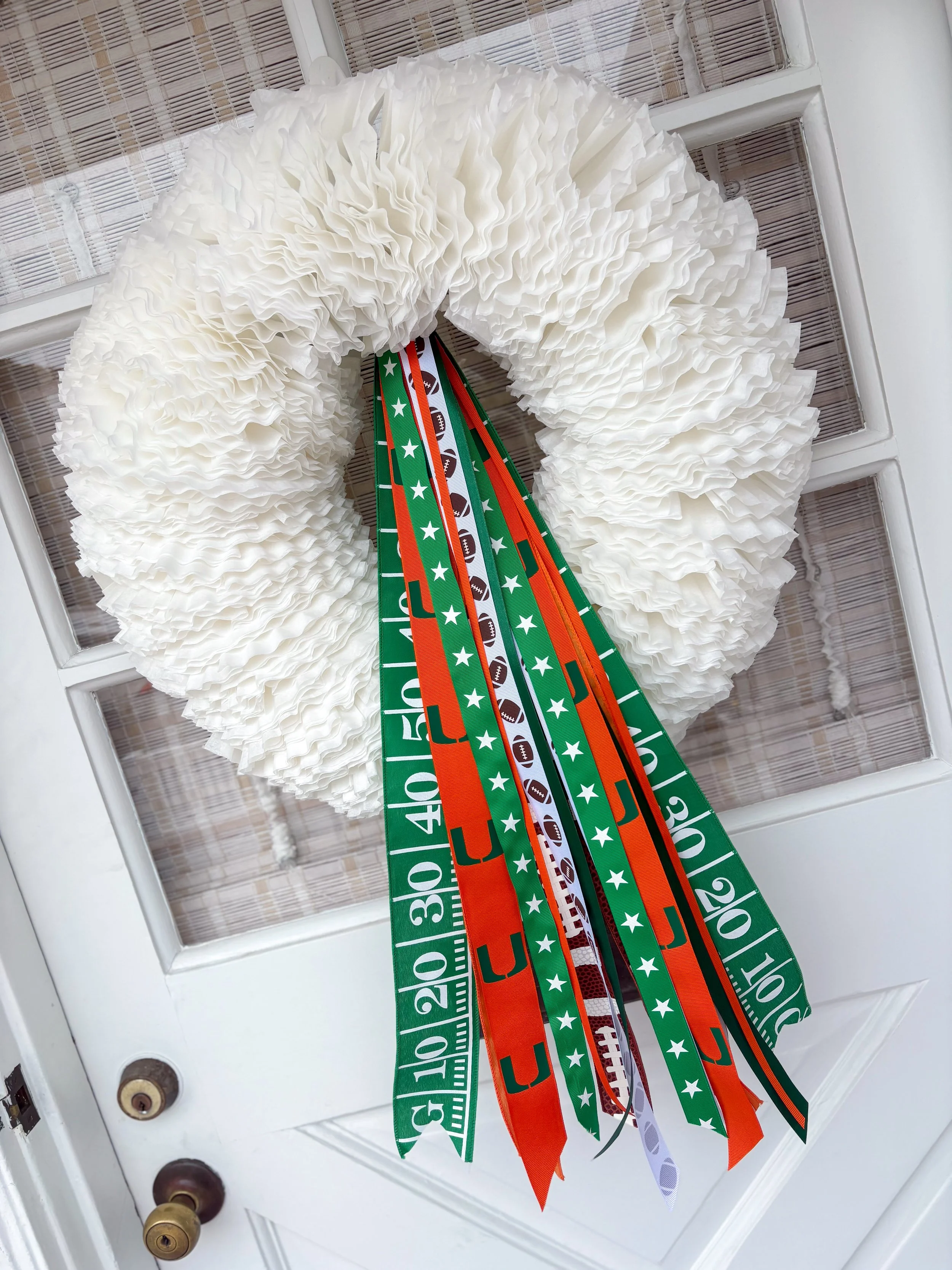 A white artificial wreath with ruffled texture hanging on a white door, decorated with green and orange ribbons featuring football and star patterns.