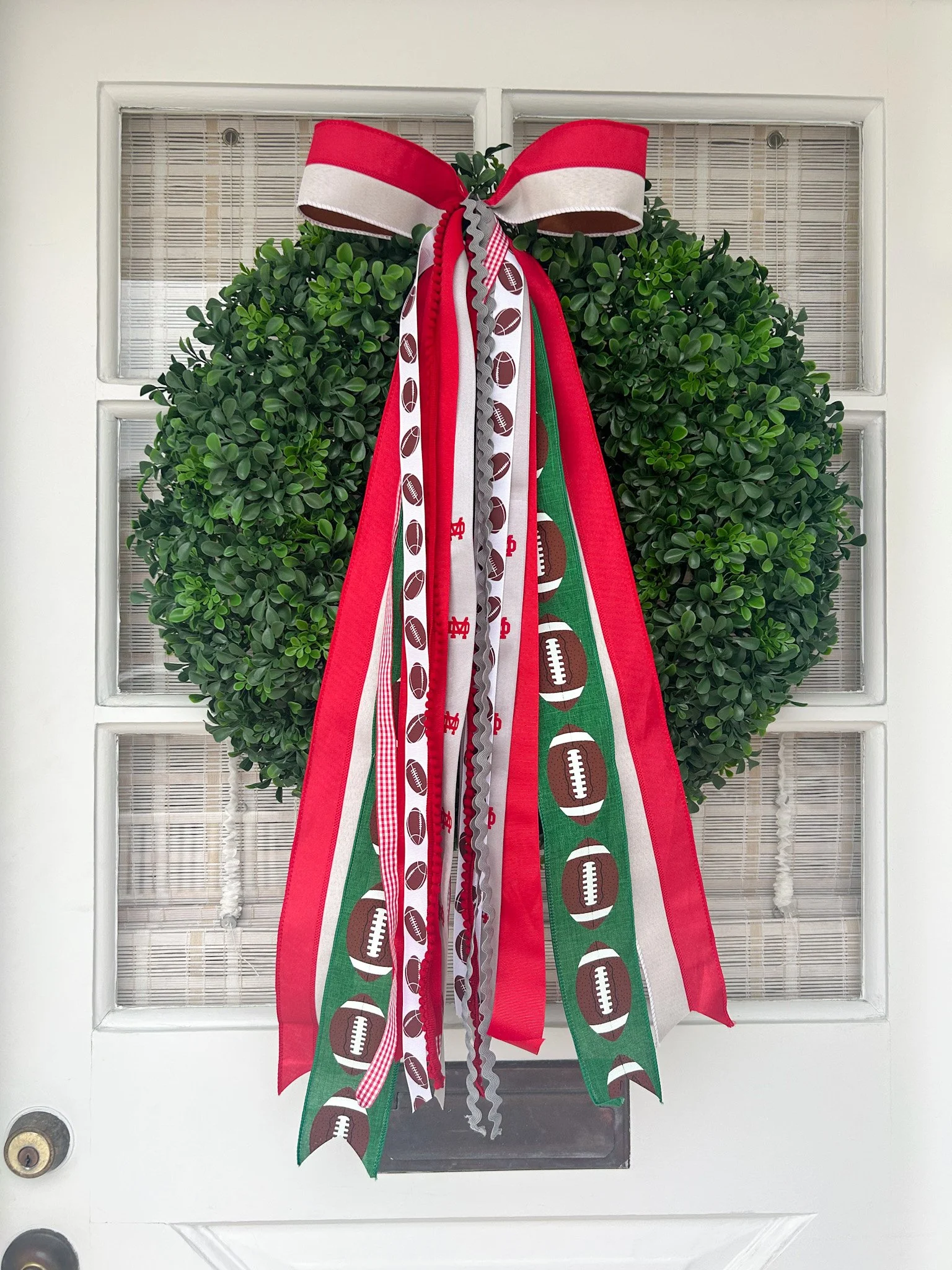 Green wreath decorated with a large red and white bow and ribbons featuring football helmets, footballs, and footballs with lattice patterns, hanging on a white door.