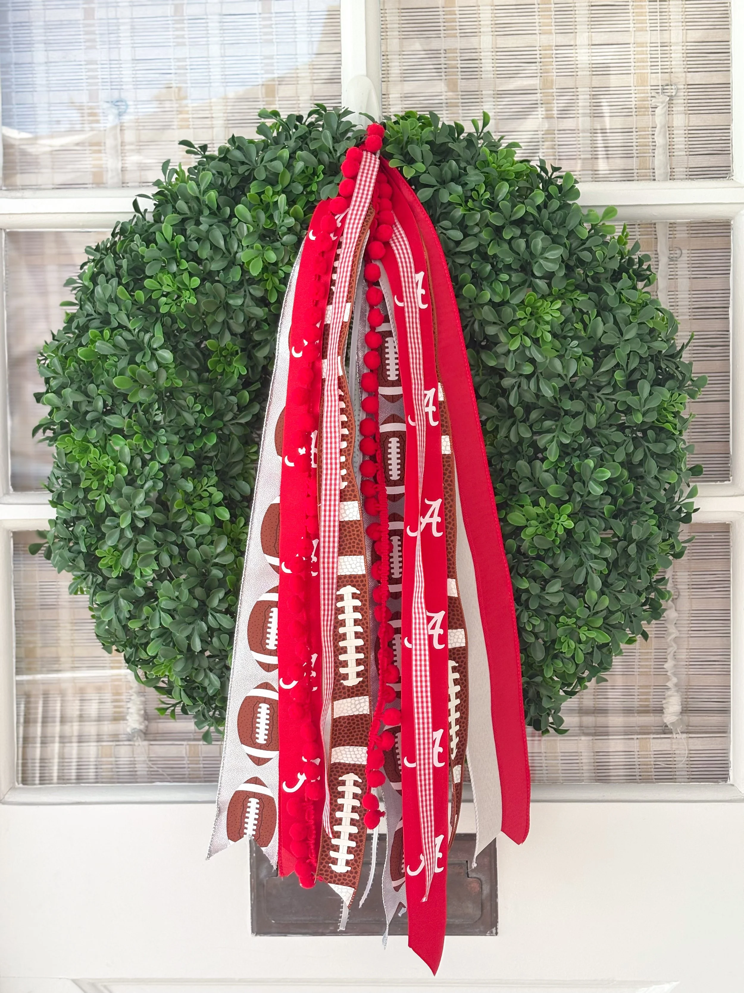 Green wreath decorated with red and white Alabama football-themed ribbons hanging on a white door with glass panels.