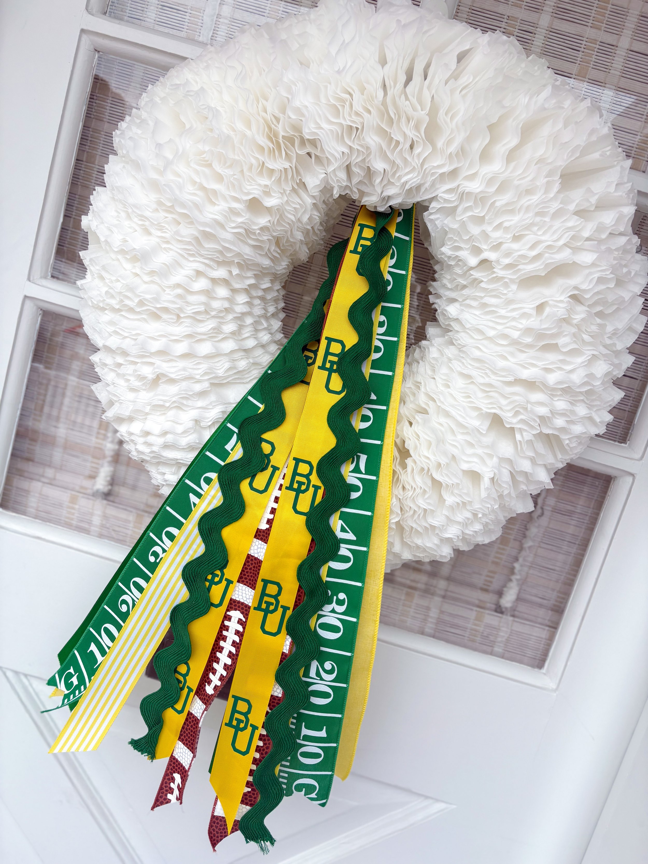 A white wreath made of coffee filters with green and yellow ribbons and a rick rack ribbon with white football patterns hanging from it, displayed on a white door.