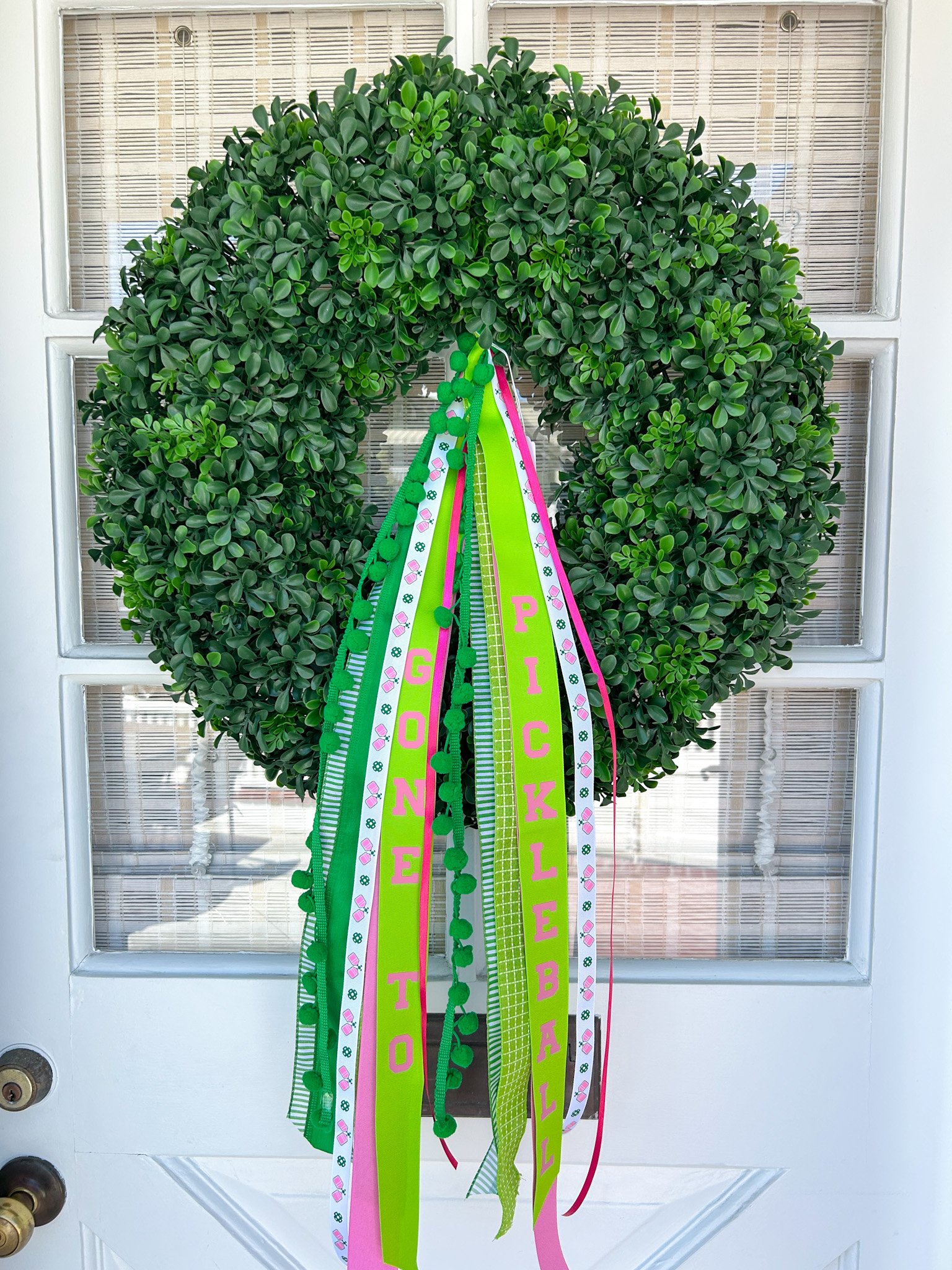 Green boxwood door wreath adorned with colorful ribbons and pins, bearing the message "GONE to PICKLEBALL".