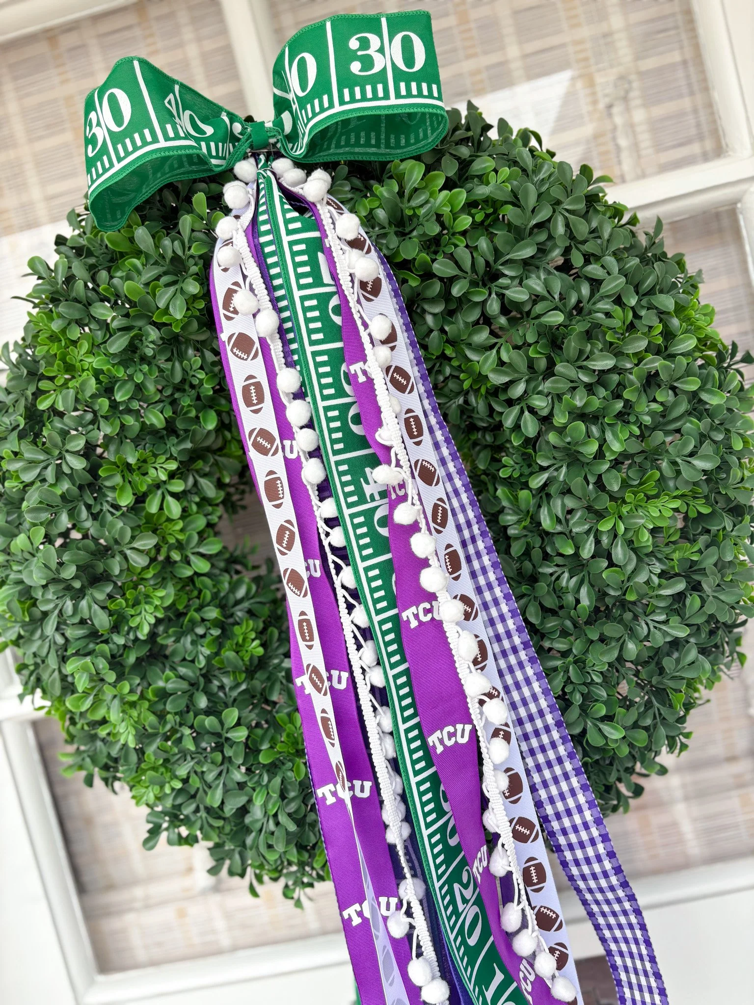 A round, green, artificial wreath decorated with colorful rally ribbons and bows in purple, white, and green, featuring texas sports team and football-themed patterns and words.
