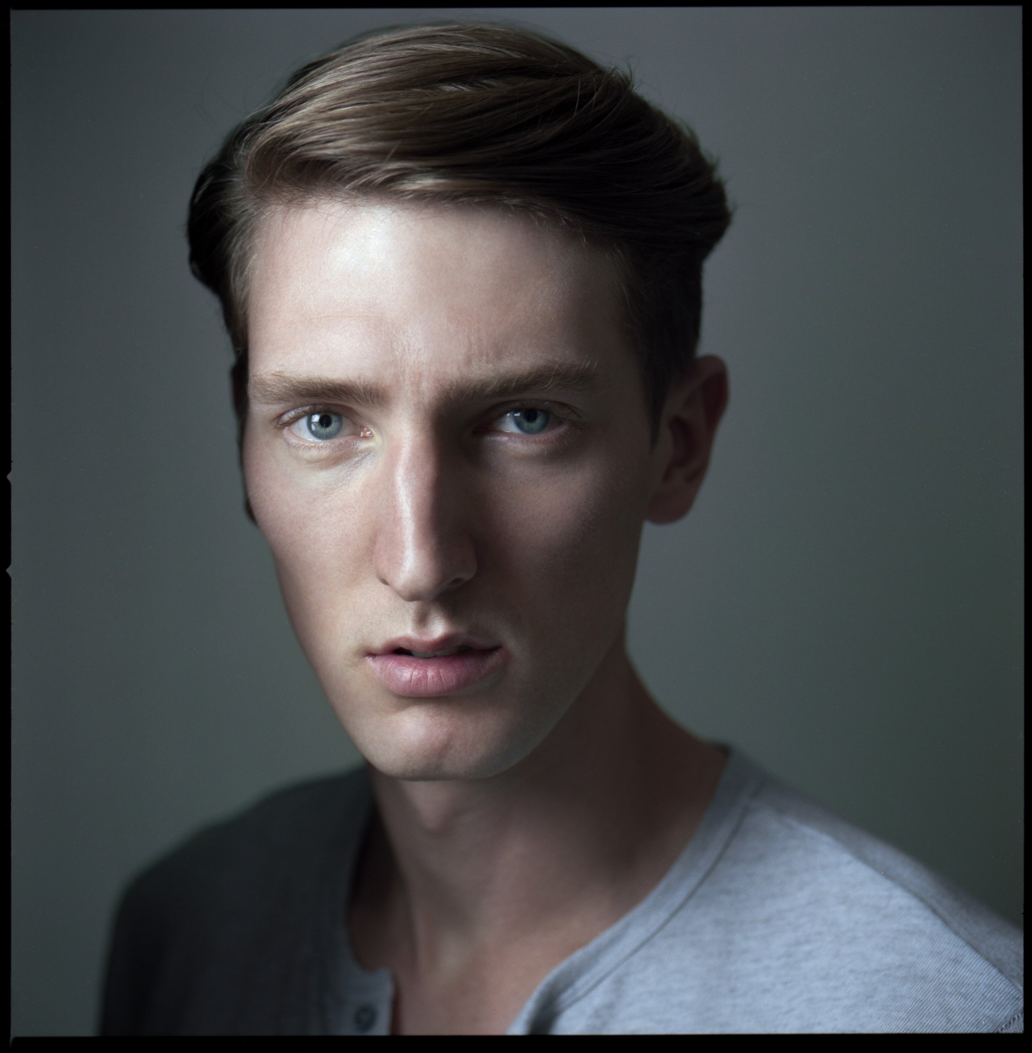 Close-up portrait of a young man with fair skin, blue eyes, and brown hair styled to the side, wearing a light grey shirt with a dark collar, against a neutral background.