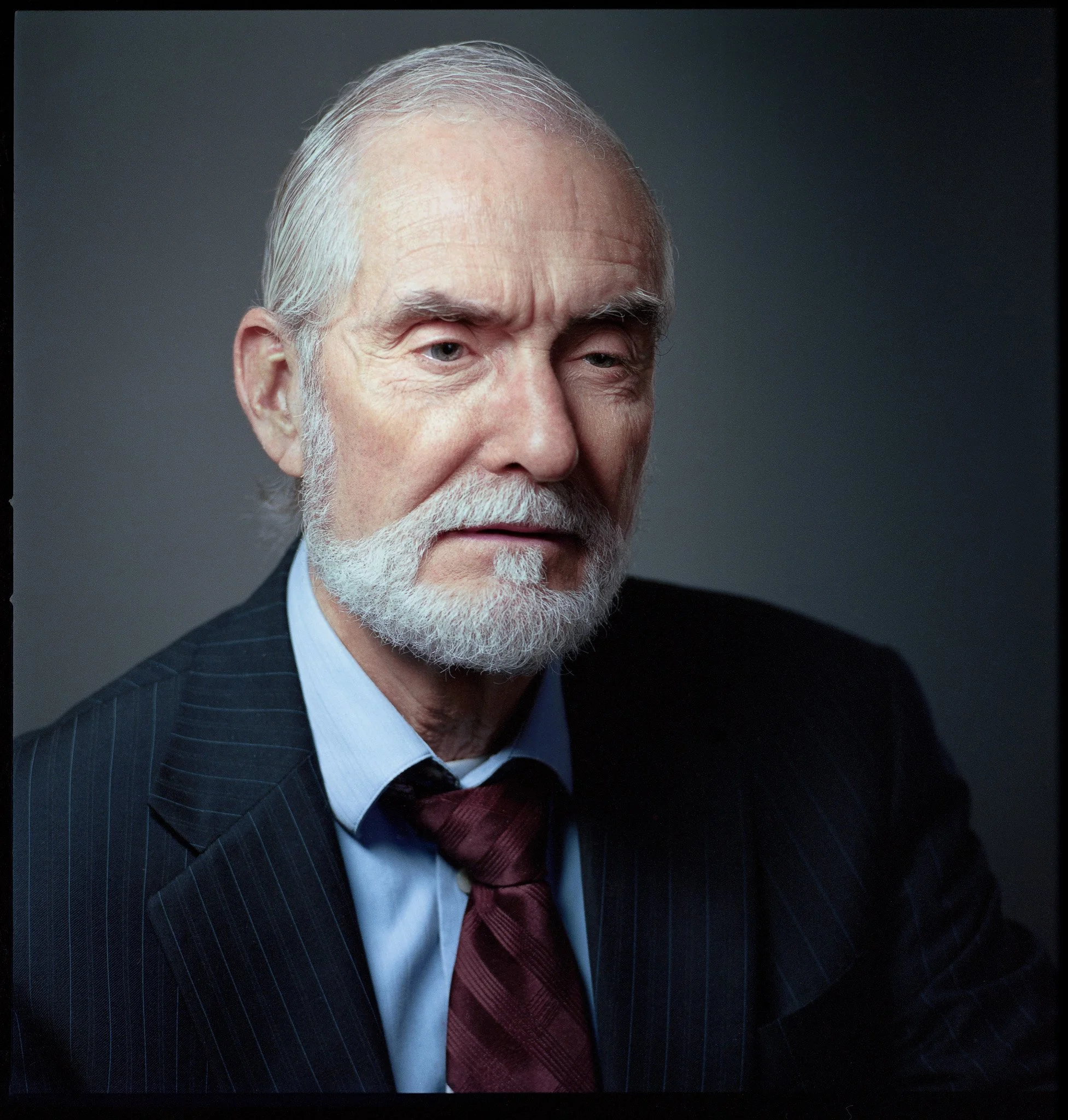 Portrait of an elderly man with white hair and beard, wearing a dark pinstripe suit, light blue dress shirt, and maroon tie, against a dark background.
