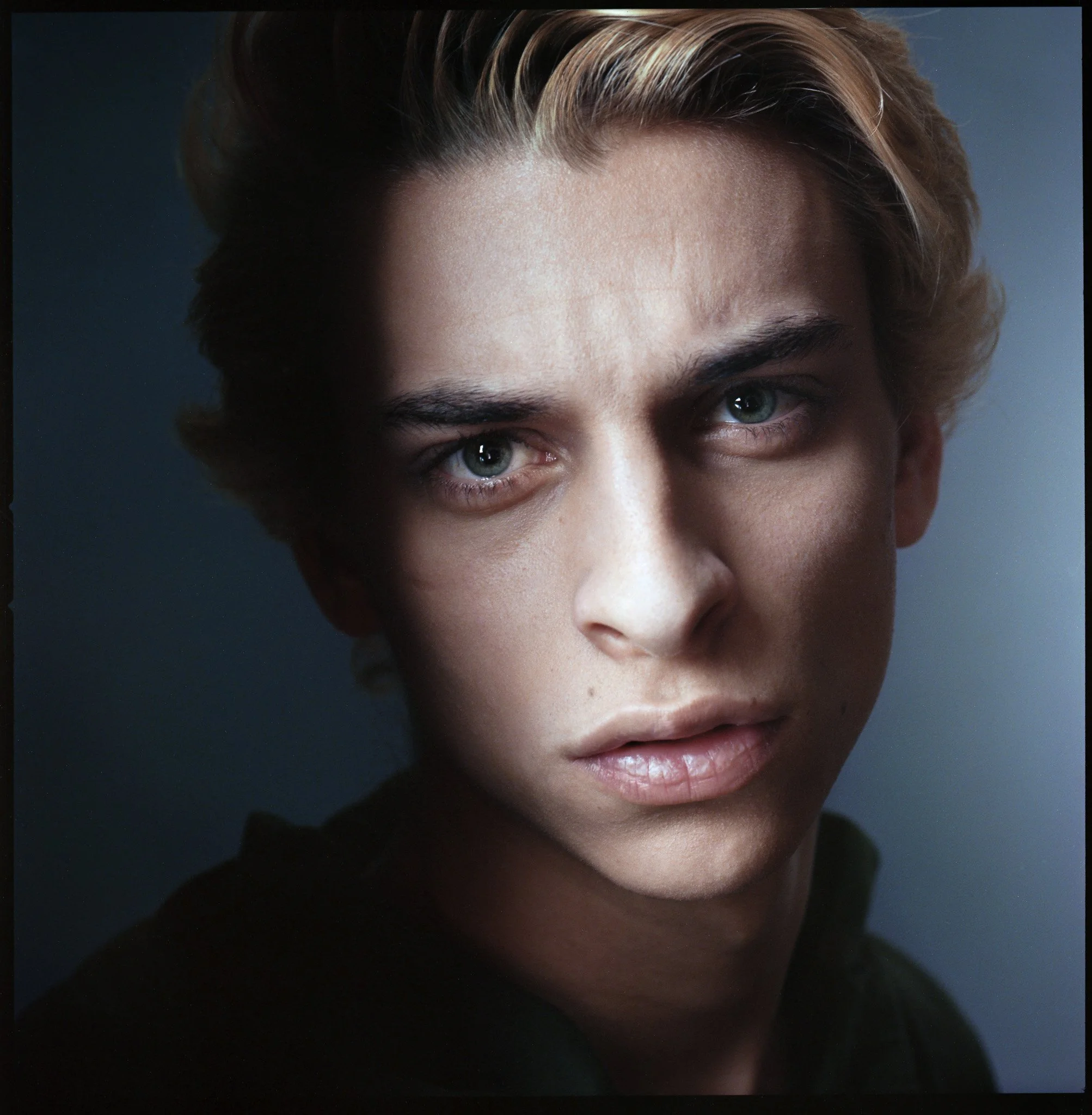 Close-up portrait of a young man with blond hair and piercing blue eyes, looking directly at the camera with a serious expression.