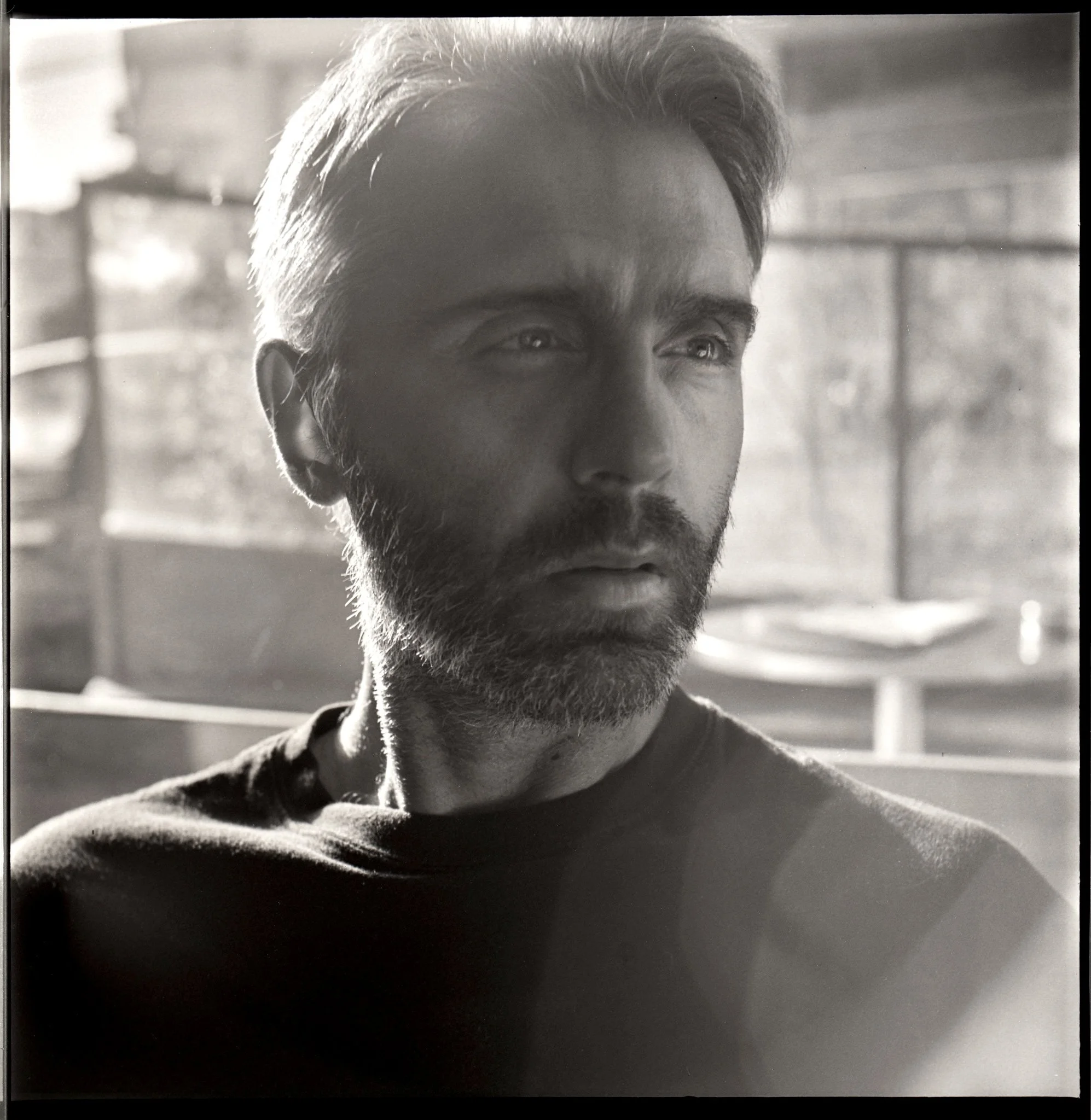 Black and white photo of a man with short hair and beard looking to the side, with sunlight coming through a window in the background.