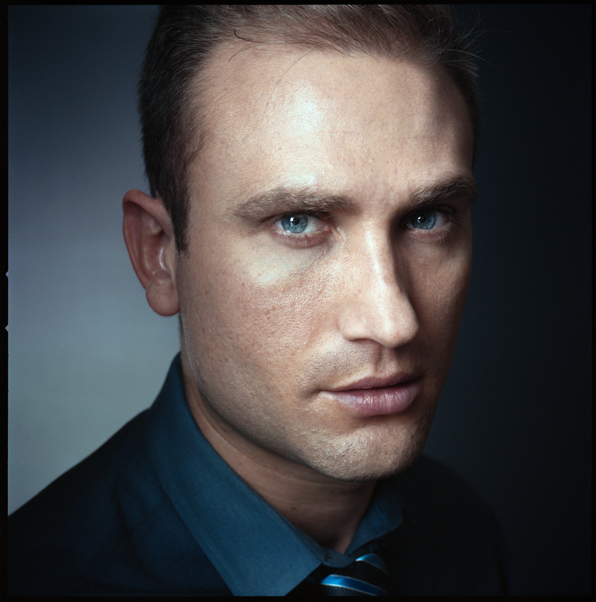 Close-up portrait of a man with blue eyes, fair skin, dark brown hair, wearing a dark suit and a striped tie, against a dark gray background.