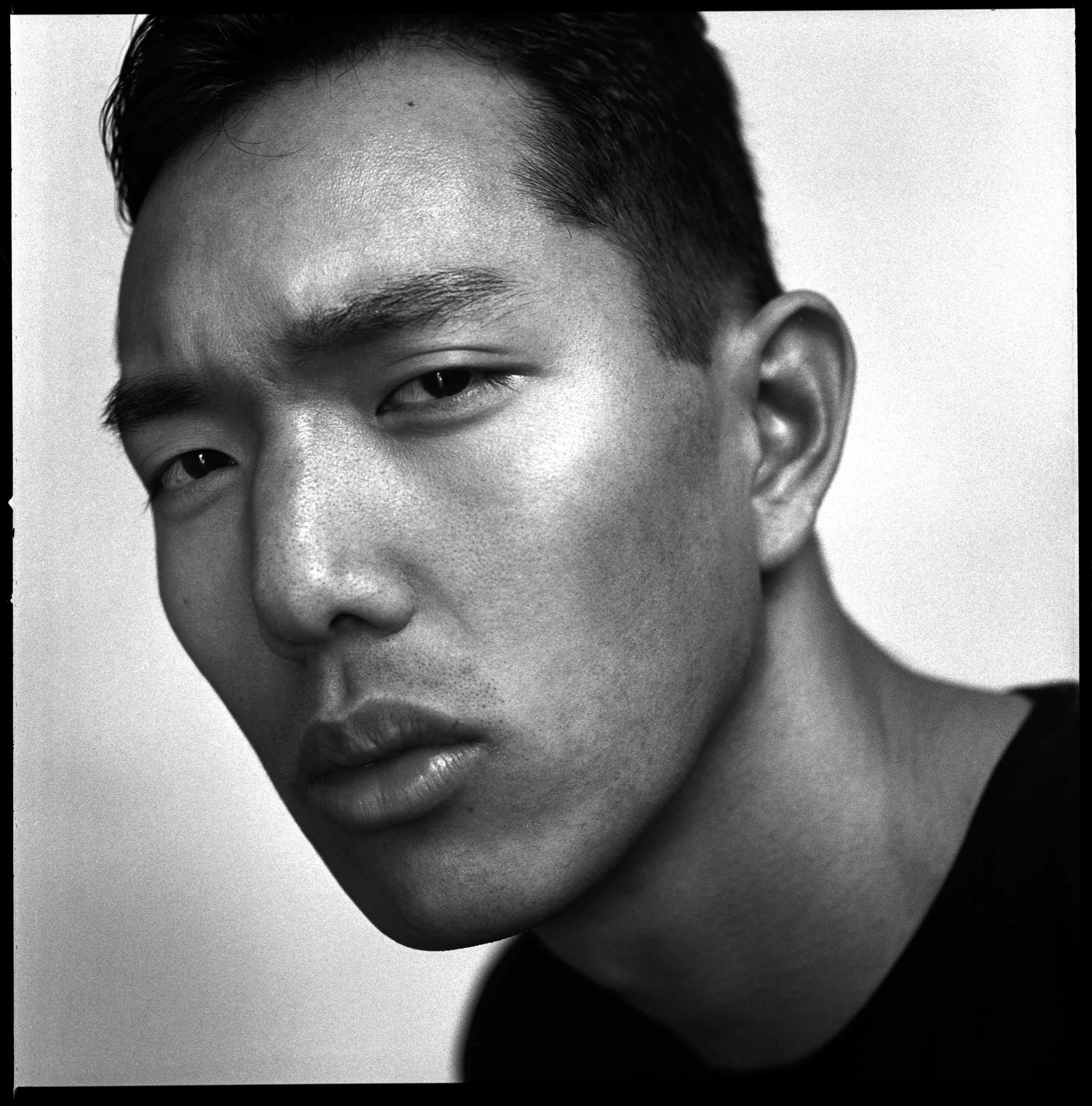 Black and white close-up portrait of a young man with short black hair, high cheekbones, and full lips, looking directly at the camera with a serious expression.