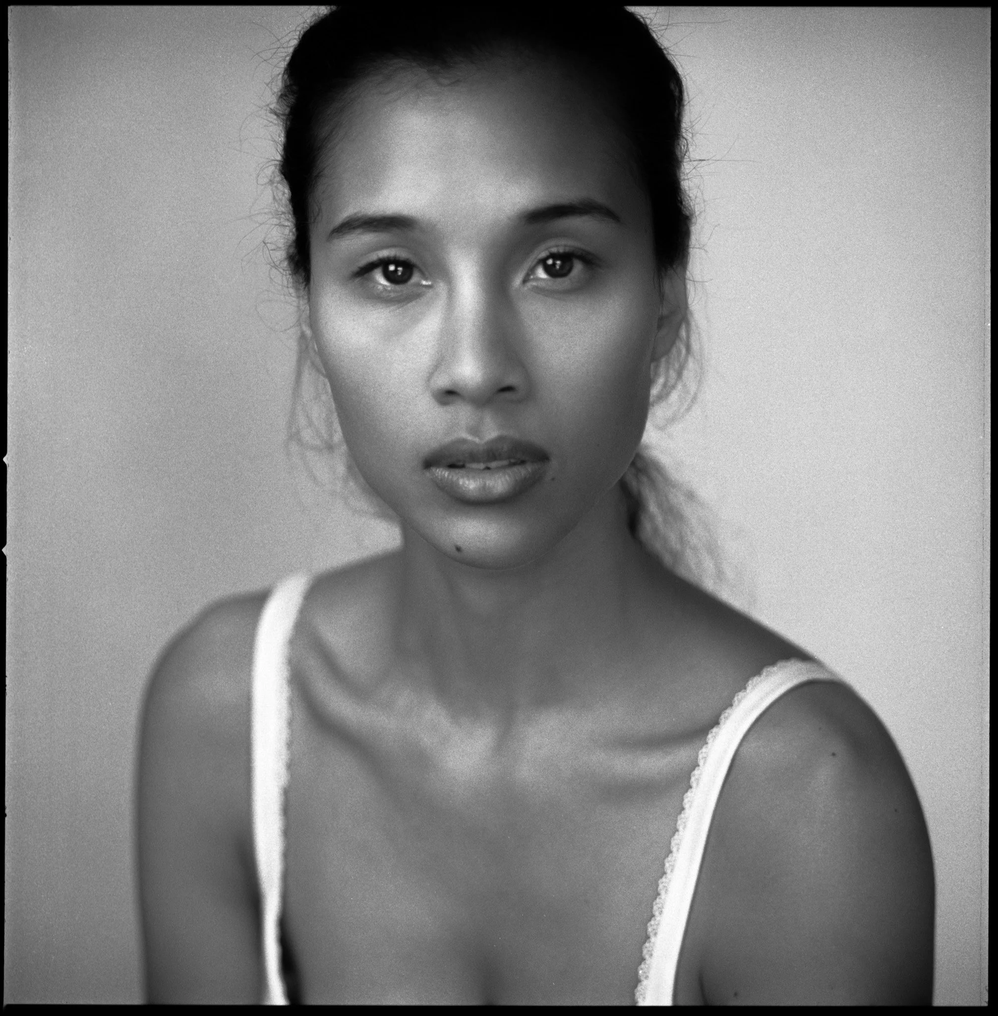 Black and white portrait of a woman with a serious expression, wearing a white tank top, looking directly at the camera.